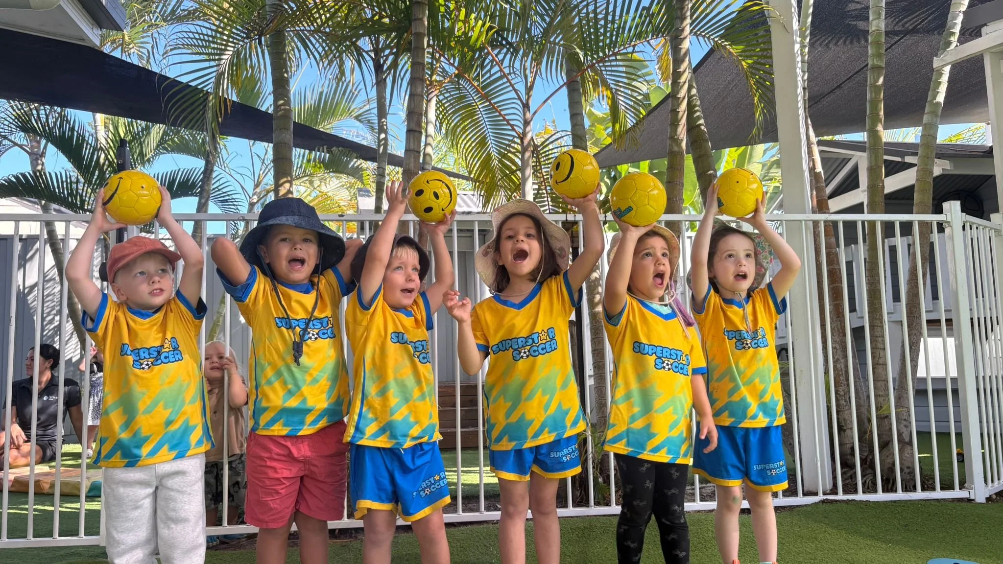 Six children in yellow and blue soccer uniforms standing outdoors, holding yellow soccer balls with smiley faces, celebrating on a grassy area with a white fence, palm trees, and a blue sky in the background.