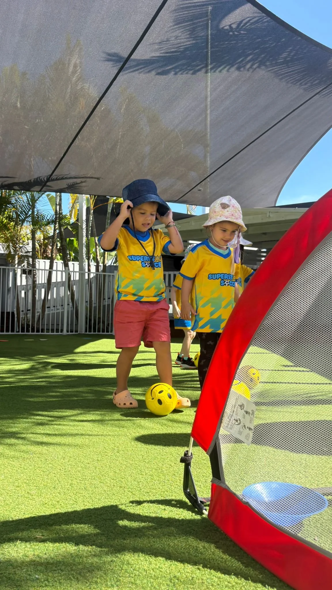 Children playing soccer outdoors on green artificial turf, wearing yellow sports jerseys, with a red and gray tent or play structure in the foreground.