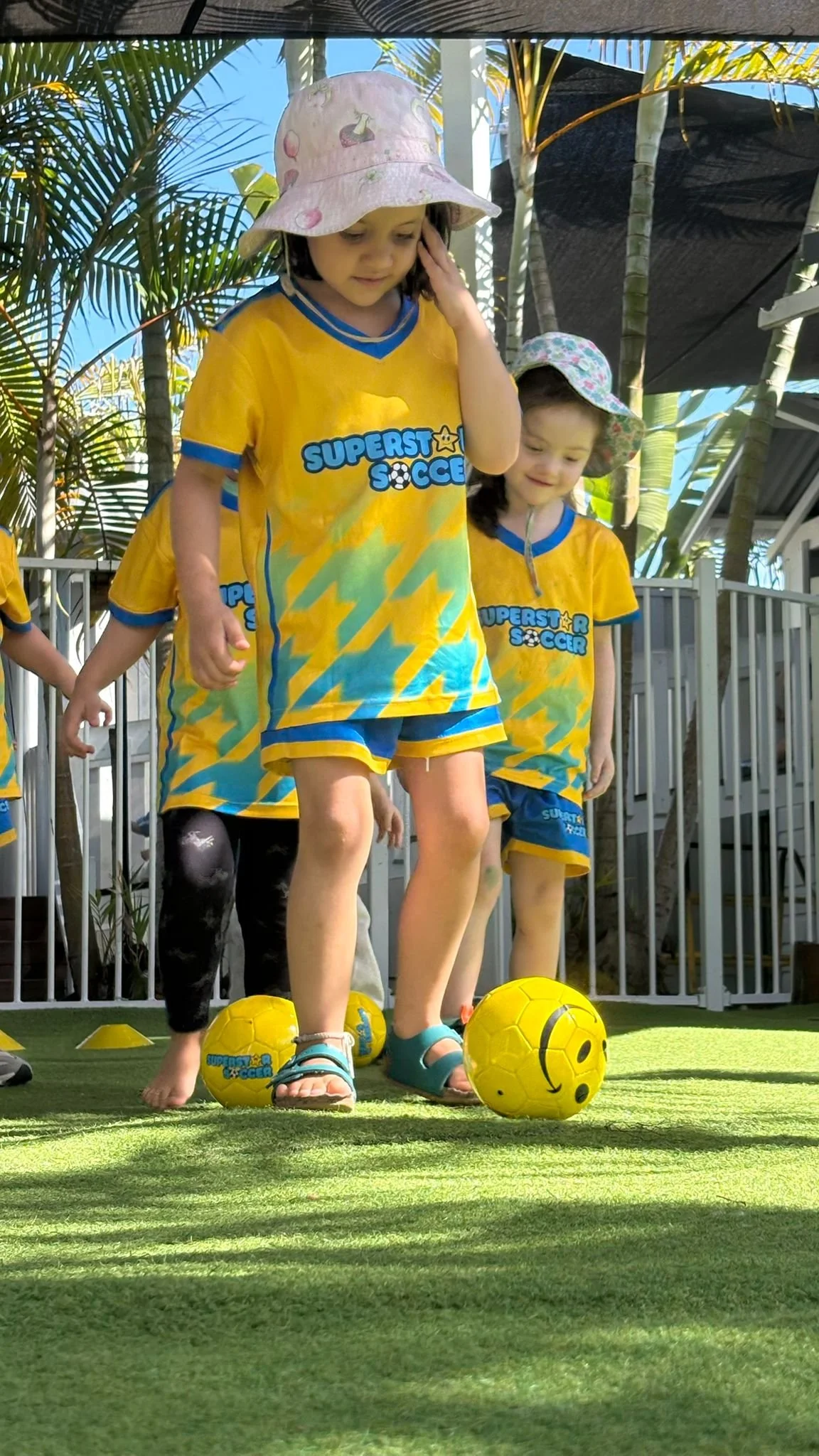 Children wearing yellow soccer jerseys and hats playing soccer on a green field with soccer balls and cones, surrounded by palm trees and a metal fence.