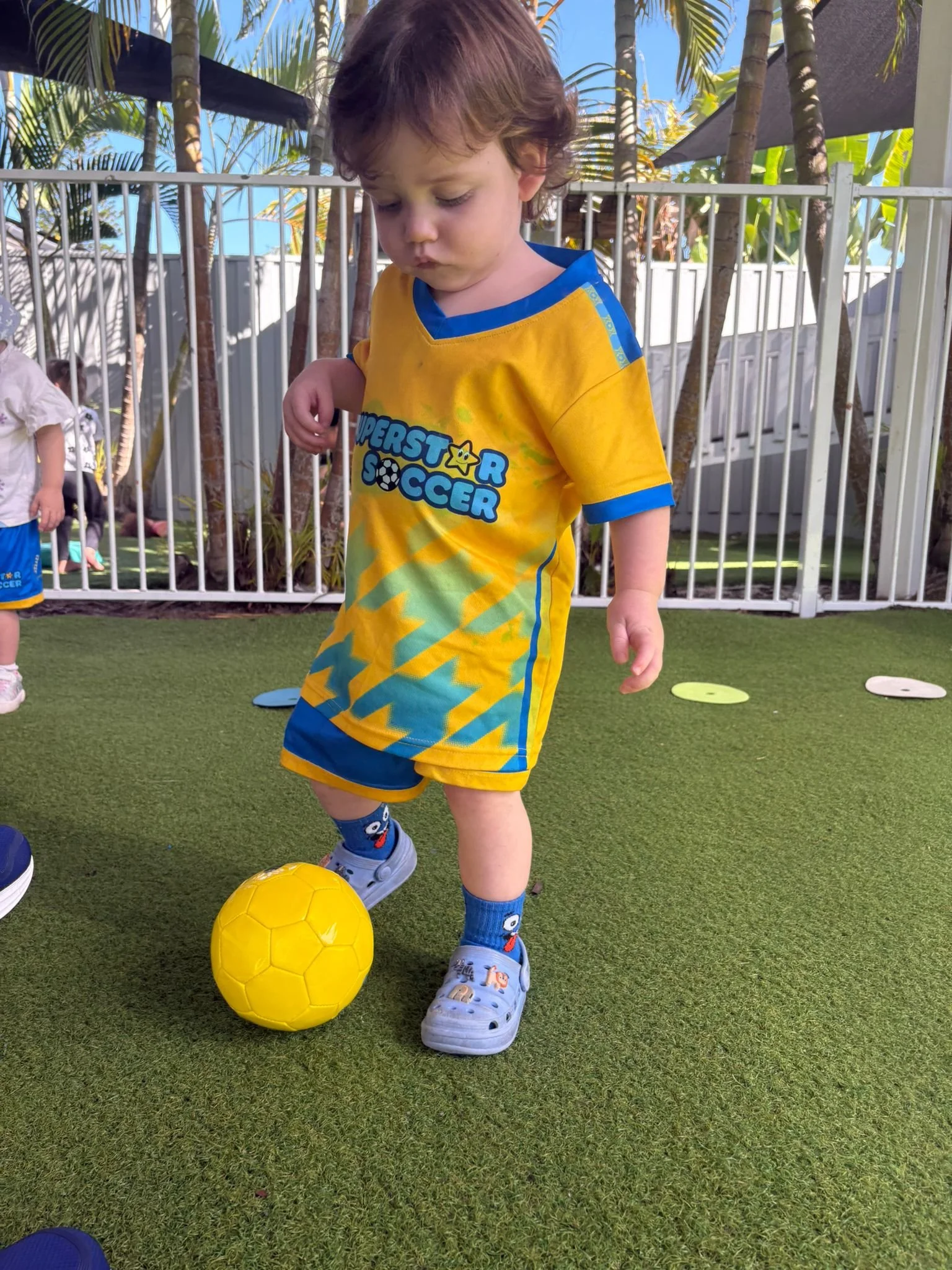 A young boy with brown curly hair playing soccer outdoors on green artificial turf. He is wearing a yellow soccer uniform with blue accents, themed 'Superstar Soccer', with colorful socks and cartoon-character sneakers. He is standing near a yellow soccer ball, with a fence, palm trees, and blue sky in the background.