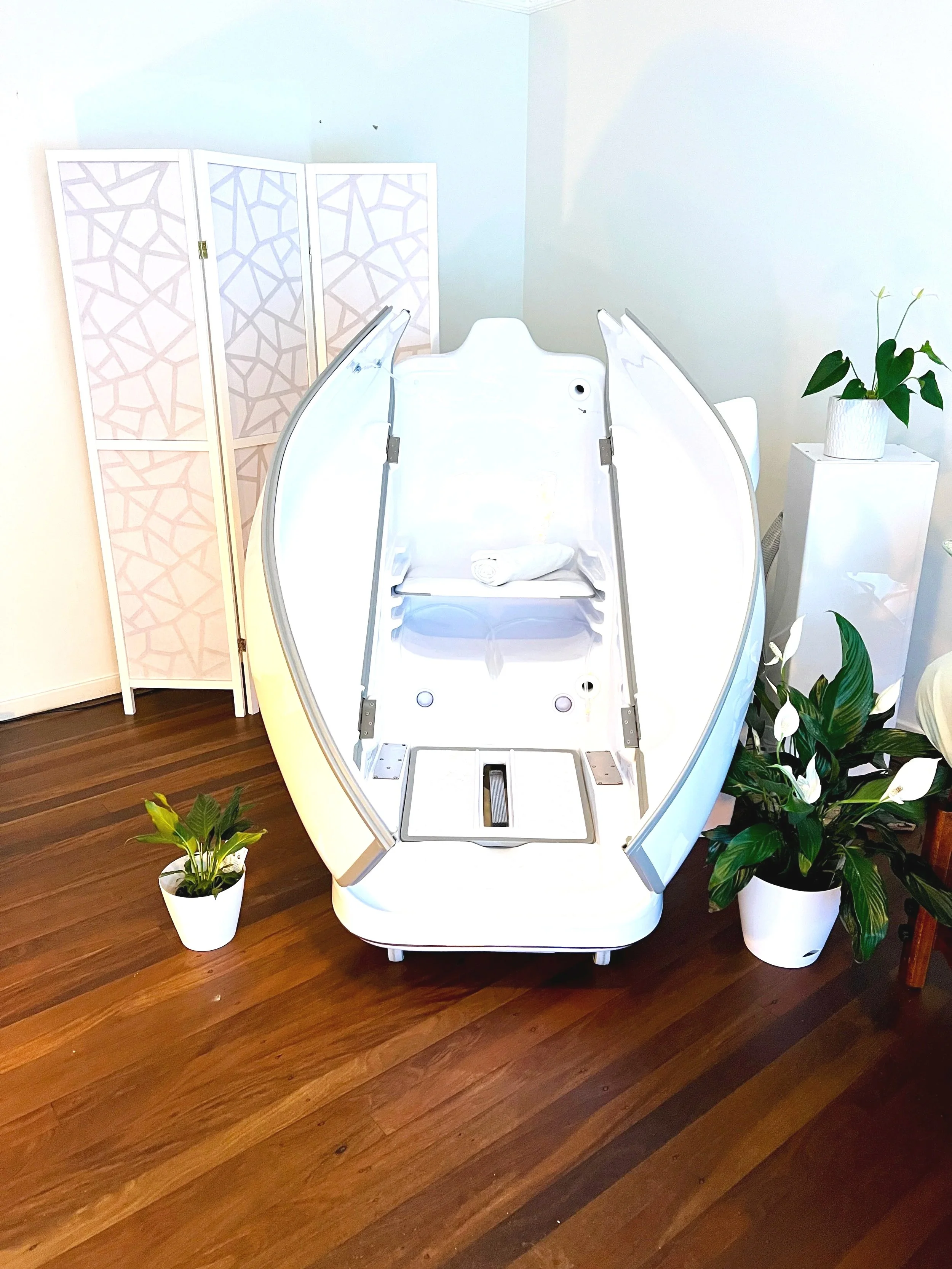 A white hyperbaric oxygen therapy chamber in a room with wooden floors. The chamber is open, showing its interior. There are potted plants on either side of the chamber, and a white privacy room divider with a geometric pattern is in the background.