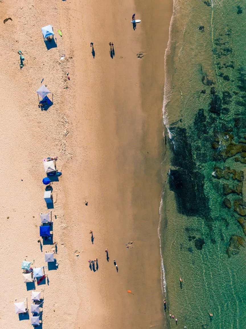 Aerial view of a sandy beach with umbrellas and people near the shoreline, transitioning into clear green water with rocks and seaweed.