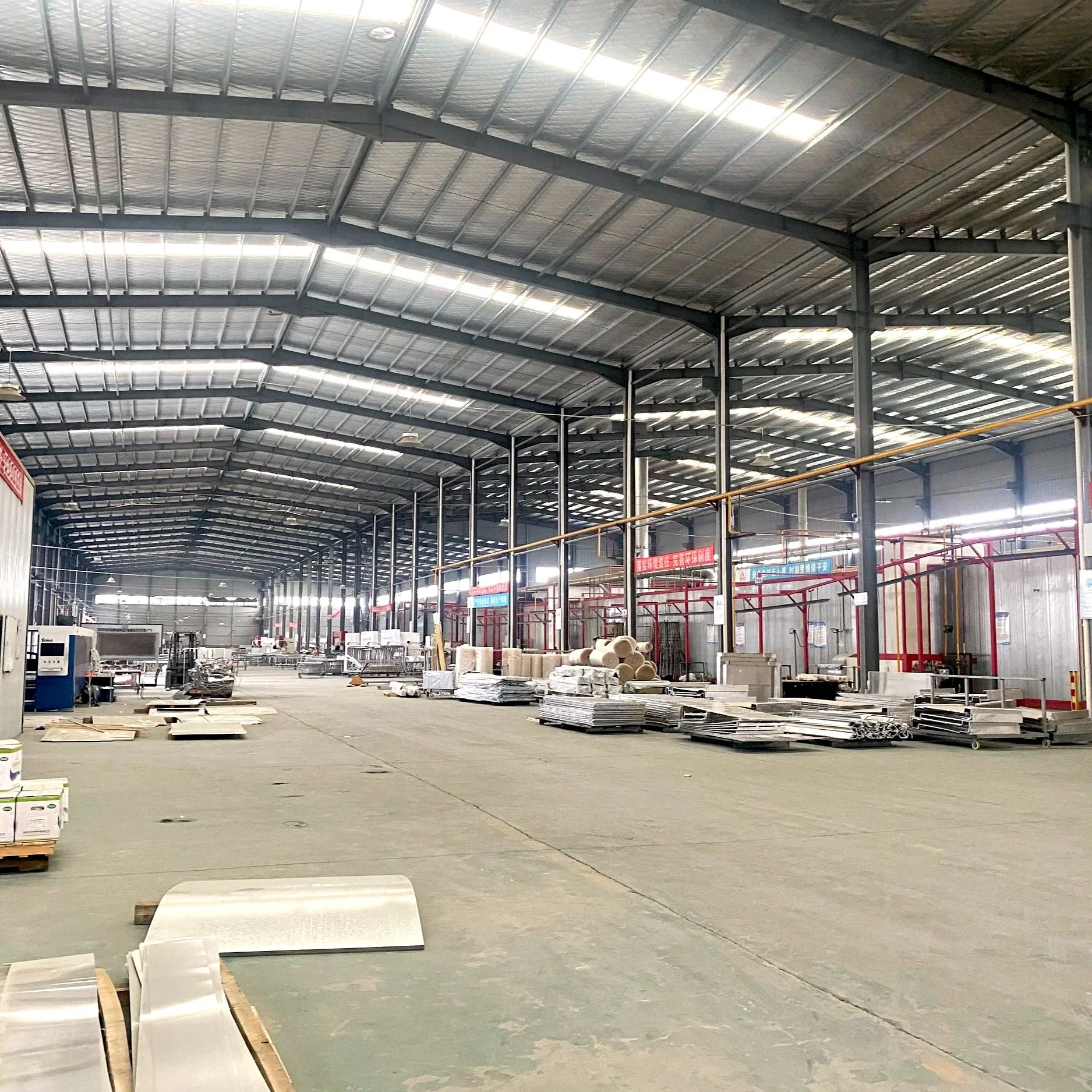 Interior of a large warehouse or factory with a high arched metal roof, pipes, and industrial equipment. There are stacked materials and machinery on the floor.