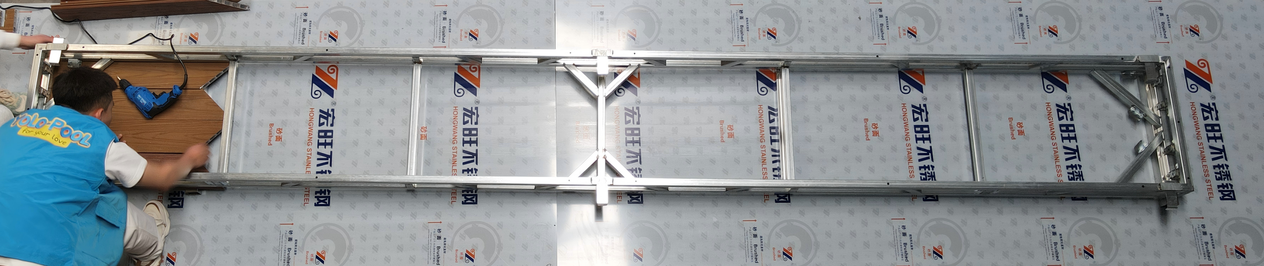 Person assembling a metal ladder on a floor covered with protective material with Chinese characters and 'Hongwang Stainless Steel' branding.