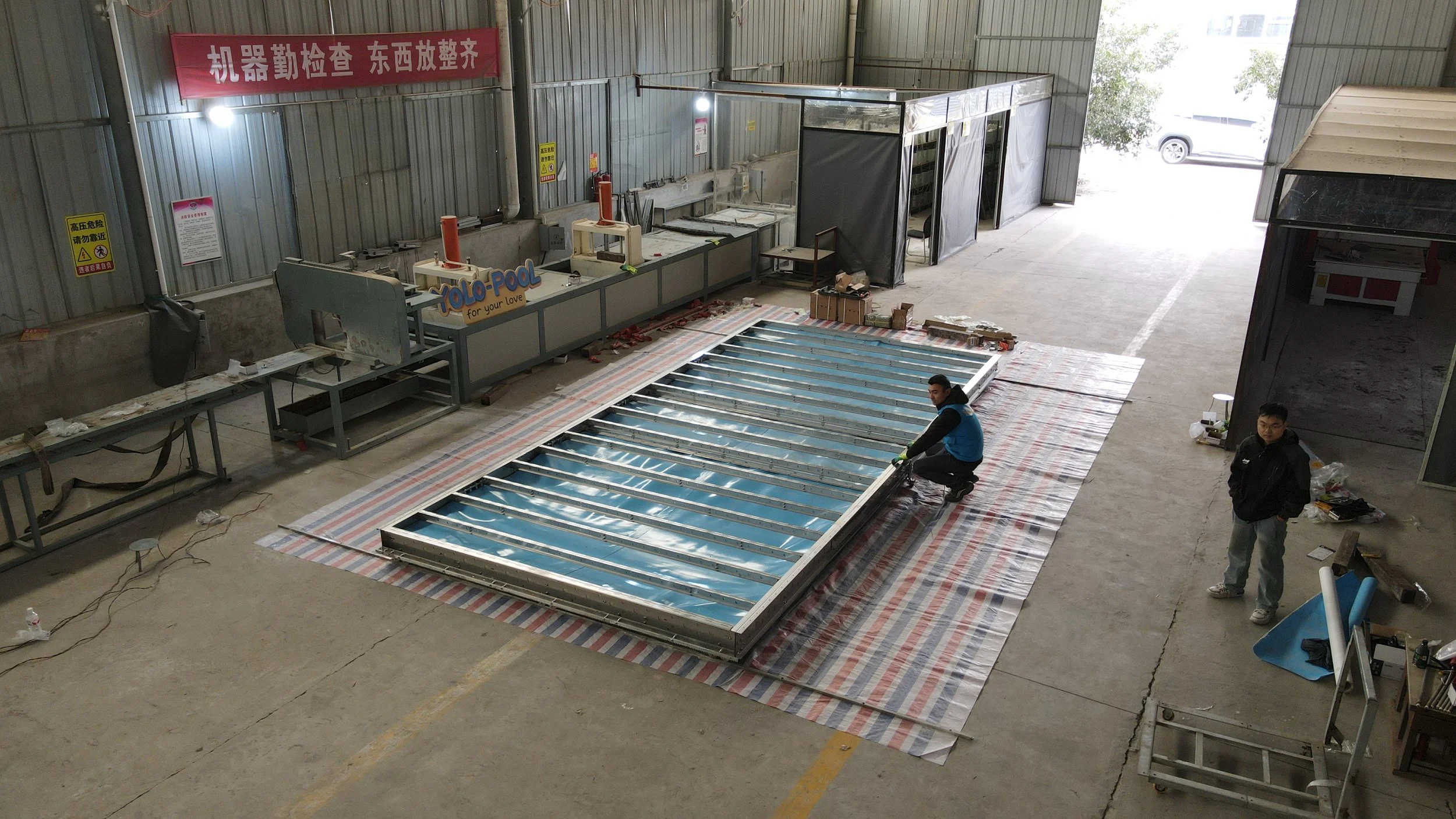 Workers assembling or repairing a large metal structure, possibly a framework or ceiling, inside a warehouse or industrial building with high ceilings and large open space. Two workers are actively working on the structure, while another stands nearby. The space contains equipment, tools, and boxes, with a striped sheet or tarp on the floor underneath the structure.