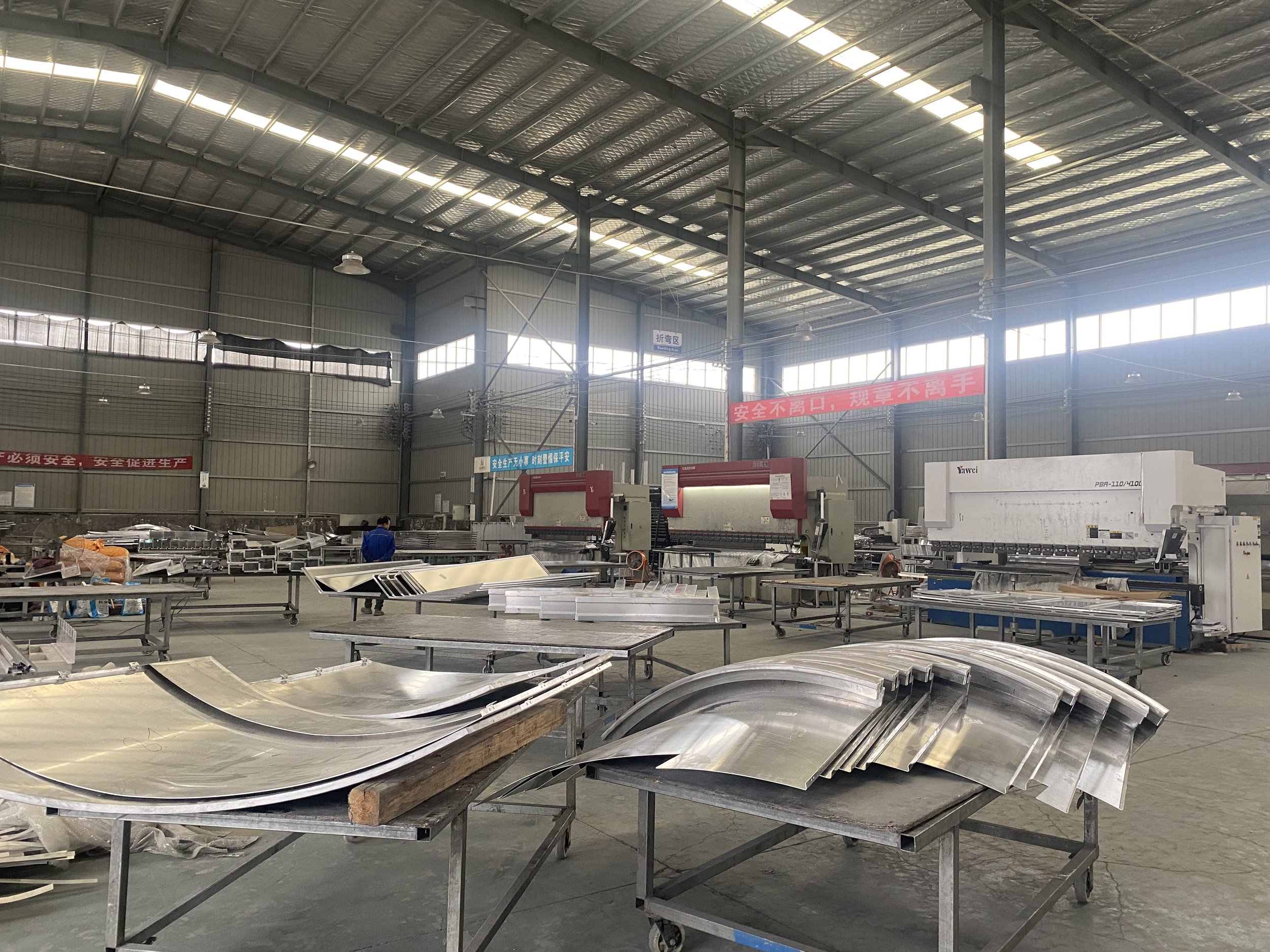 Interior of a factory or warehouse with metal roofing and high windows, containing metal sheets and equipment used for manufacturing or construction.