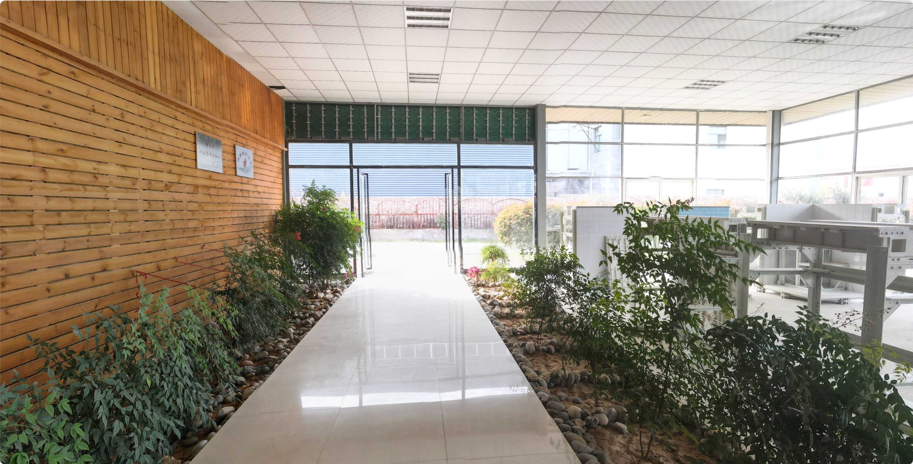 Indoor space with plants along a lined walkway leading to glass double doors, with windows on the right showing sunlight and a garden outside, and wood paneling on the left wall.