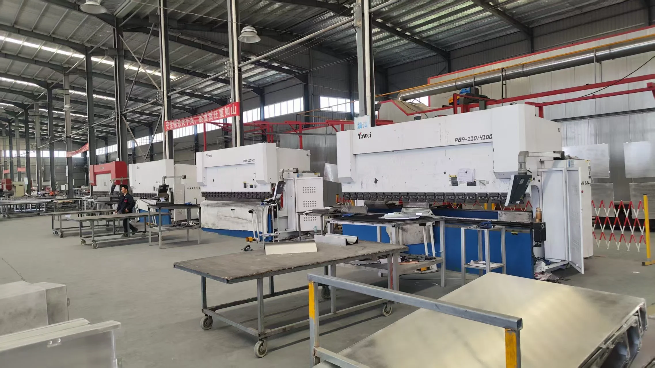 Industrial factory with multiple large CNC machines and work tables, some workers nearby, metal construction, high ceiling, bright lighting.