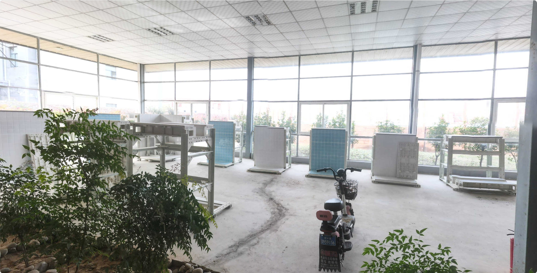 Interior of a bright room with large windows, unfinished floor, plants in the foreground, and several racks or shelves along the window wall, with a small electric bike parked near the plants.