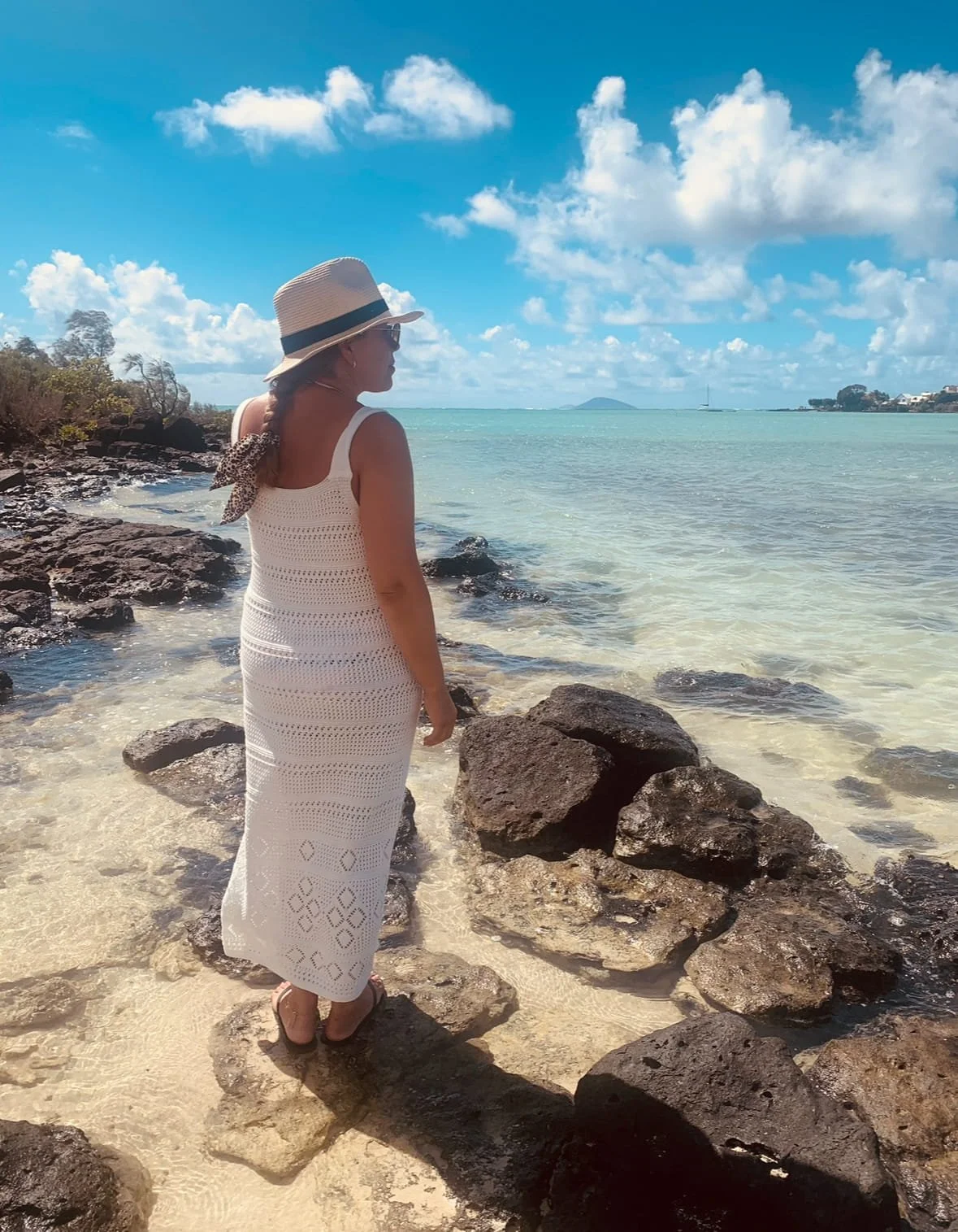 A woman wearing a white crocheted dress, a wide-brimmed hat, and sandals stands on rocks by the ocean, looking at the water with a cloudy blue sky overhead.