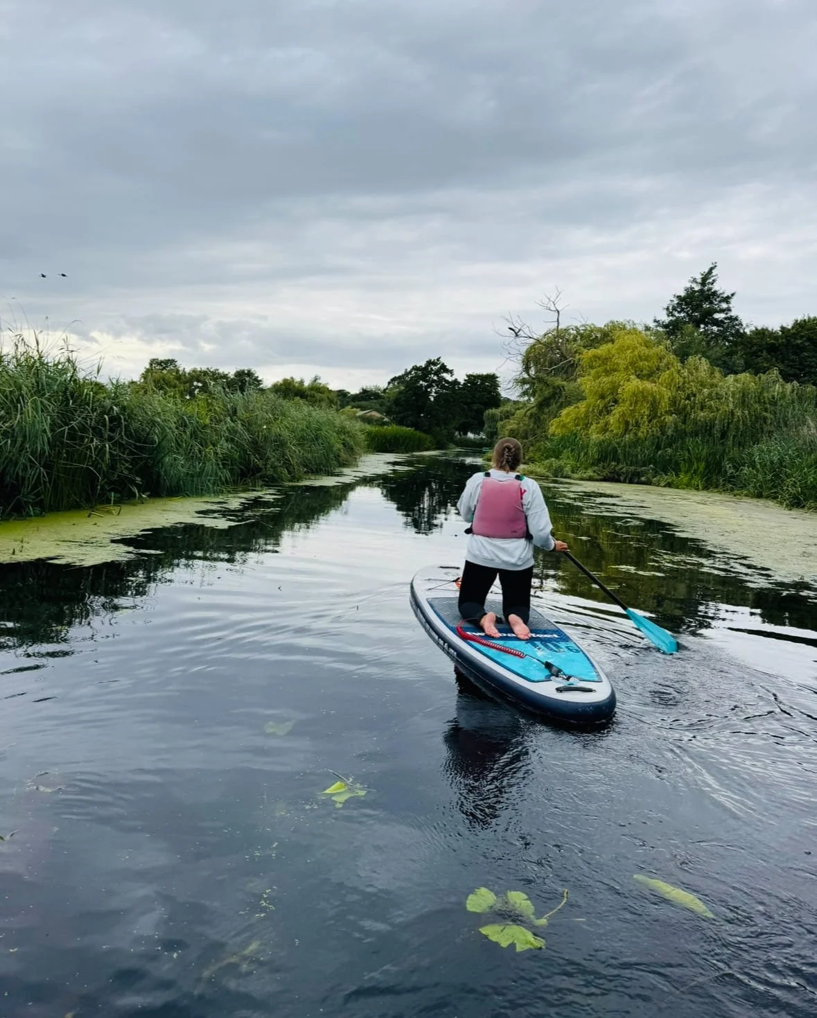 Person paddleboarding on a calm river surrounded by green tall grass and trees on a cloudy day.