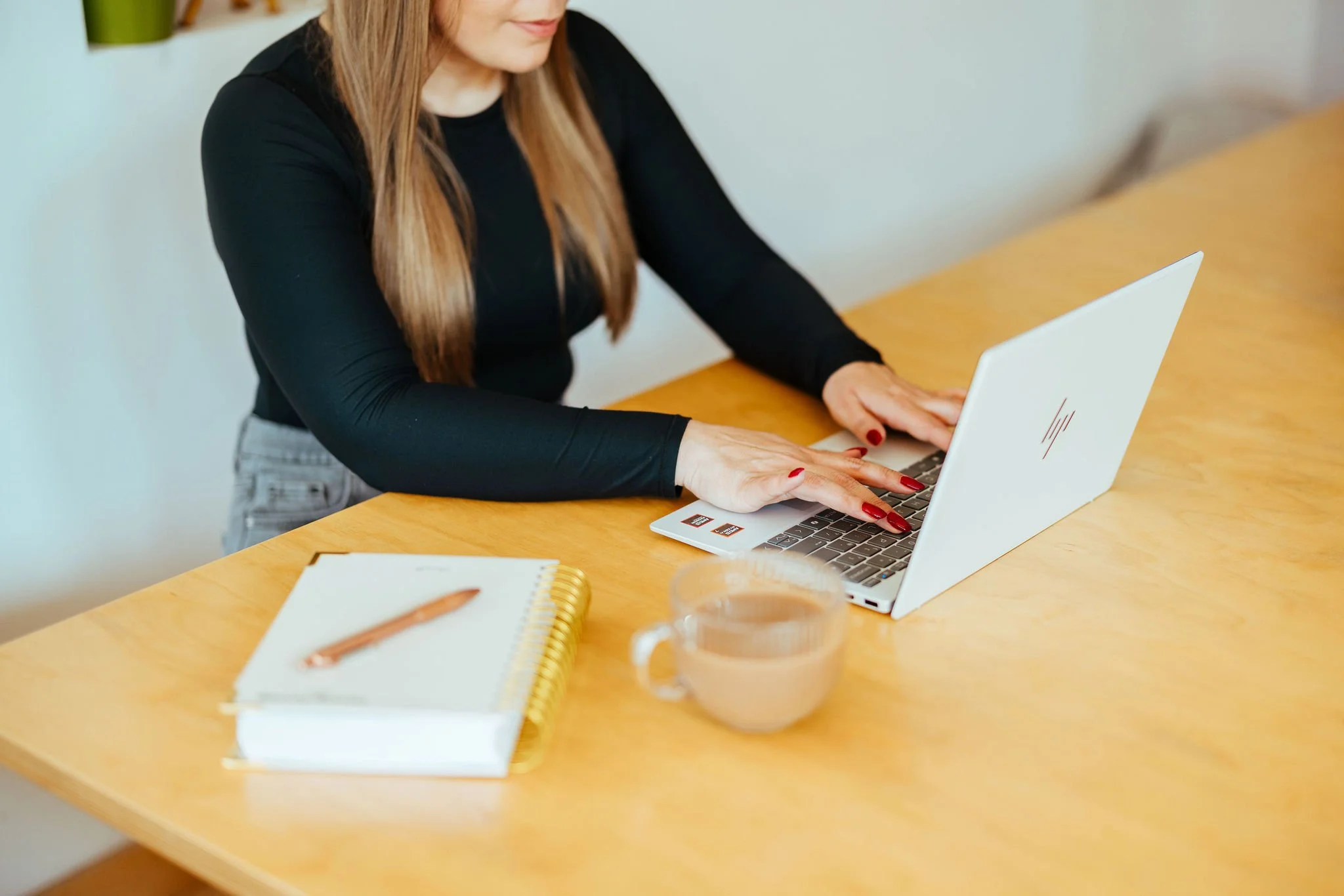 A woman with long hair working on a silver laptop at a wooden table, with a notebook, pen, and a glass cup of coffee nearby.