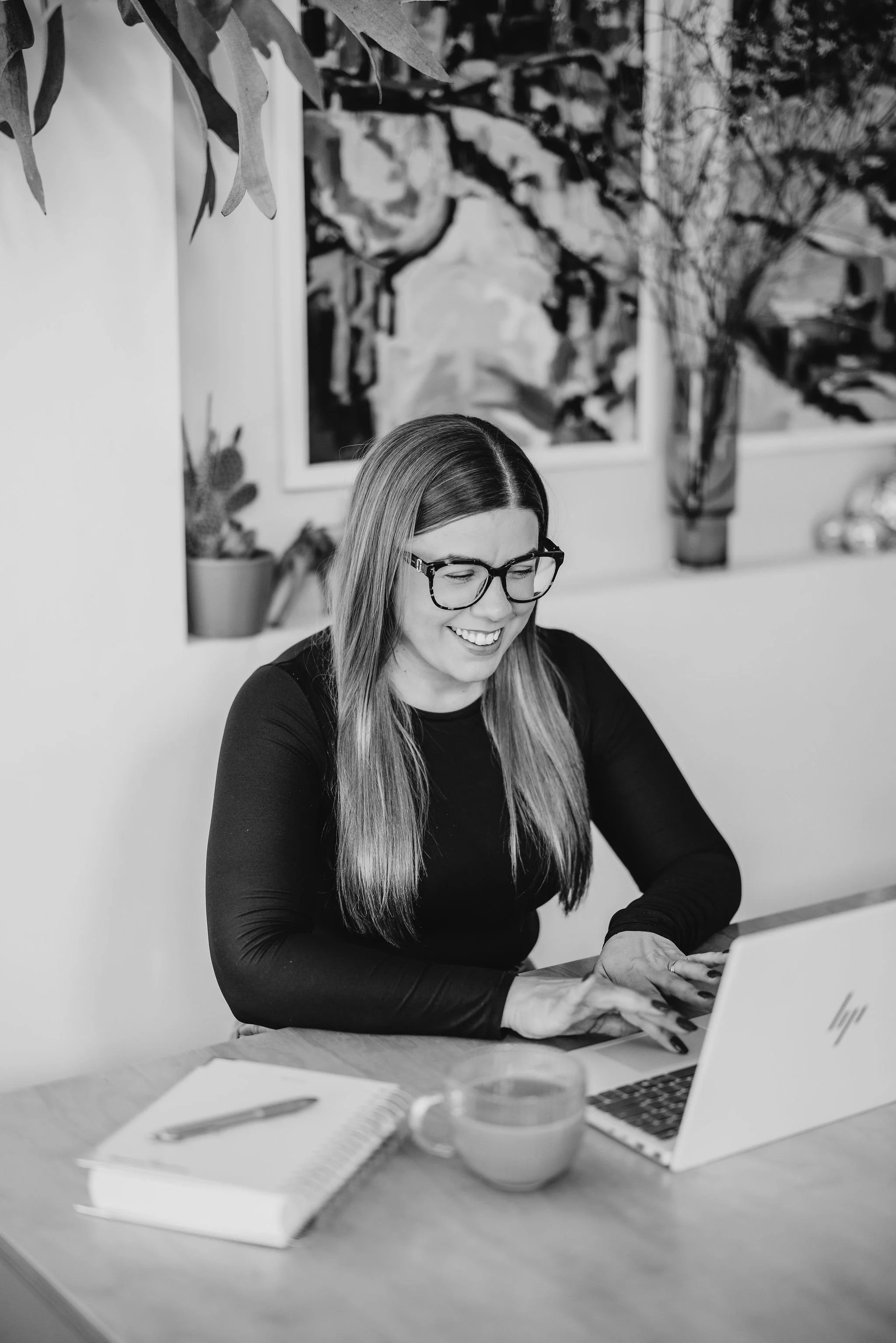 A woman with glasses smiling and working on a laptop at a desk, with a notebook, pen, and cup nearby, in a room with artwork and plants on the wall.