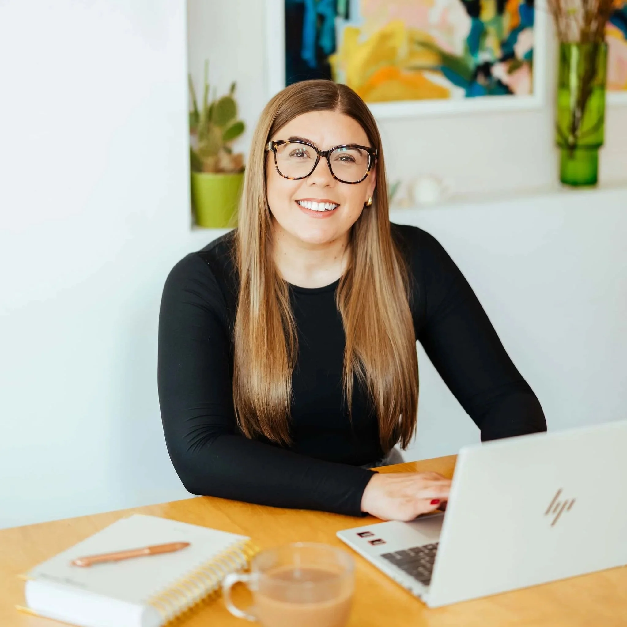 A woman with long brown hair, wearing glasses and a black shirt, smiling at her laptop at a wooden table in a bright room with colorful artwork and houseplants in the background.