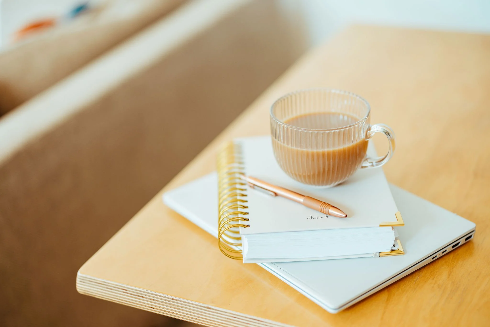 A clear glass cup filled with coffee or tea placed on top of a closed white notebook and a spiral-bound planner, with a rose gold pen resting on the notebook, all on a wooden table.