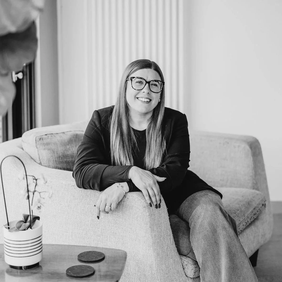 A Black and white photo of a woman sitting on a sofa, smiling and wearing glasses.