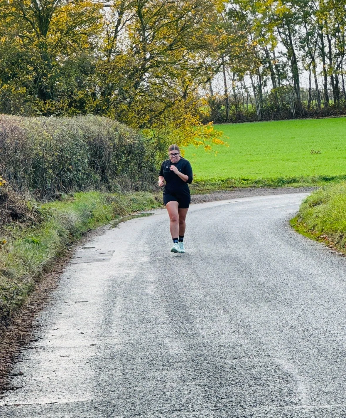 A woman jogging on a winding rural road surrounded by greenery and trees with changing autumn leaves.