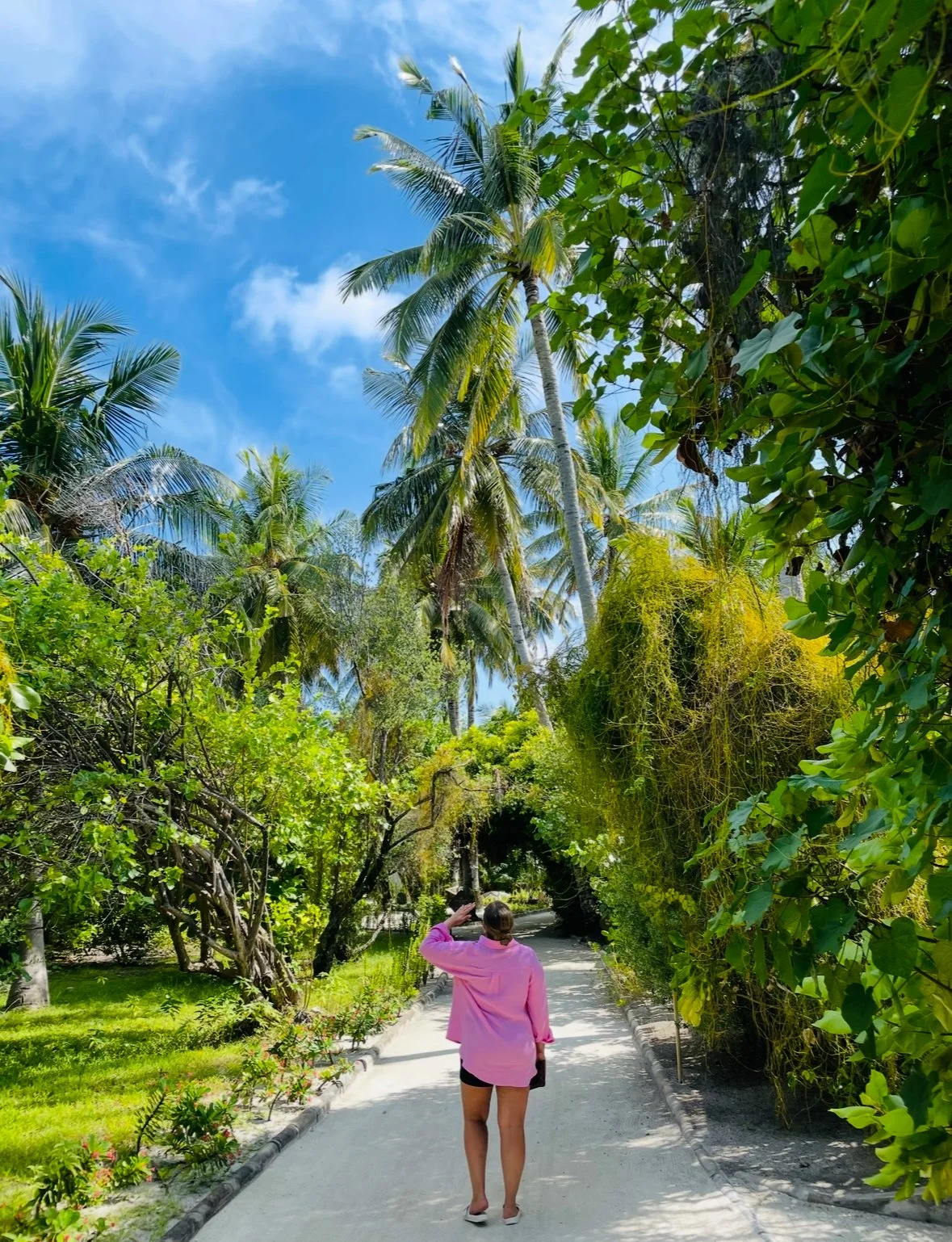 A woman in a pink top and black shorts walks on a sandy path through lush tropical greenery and tall palm trees under a partly cloudy blue sky.