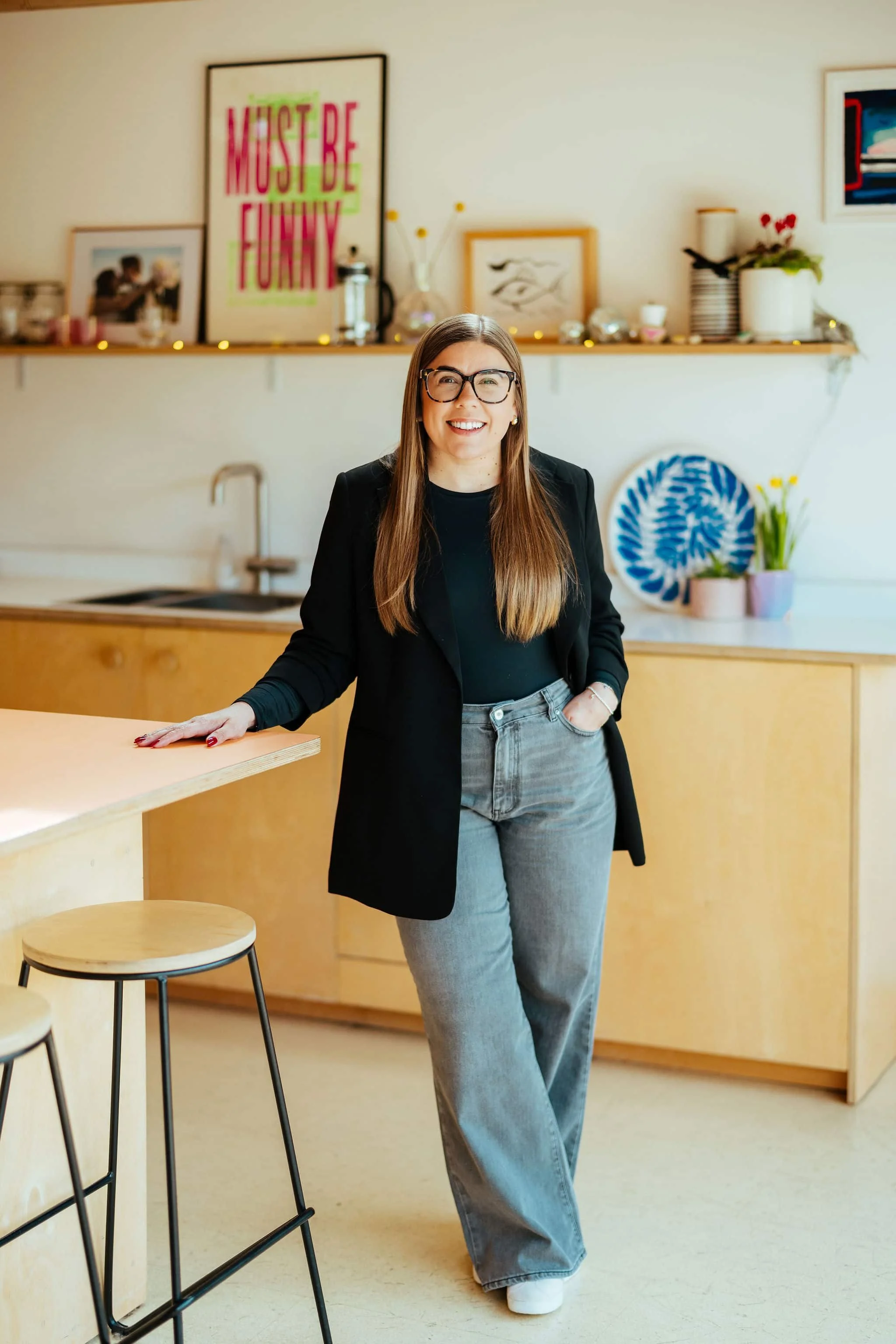 A smiling woman with long brown hair and glasses, wearing a black blazer and gray jeans, standing in a modern kitchen with light wood cabinets and decorative items in the background.