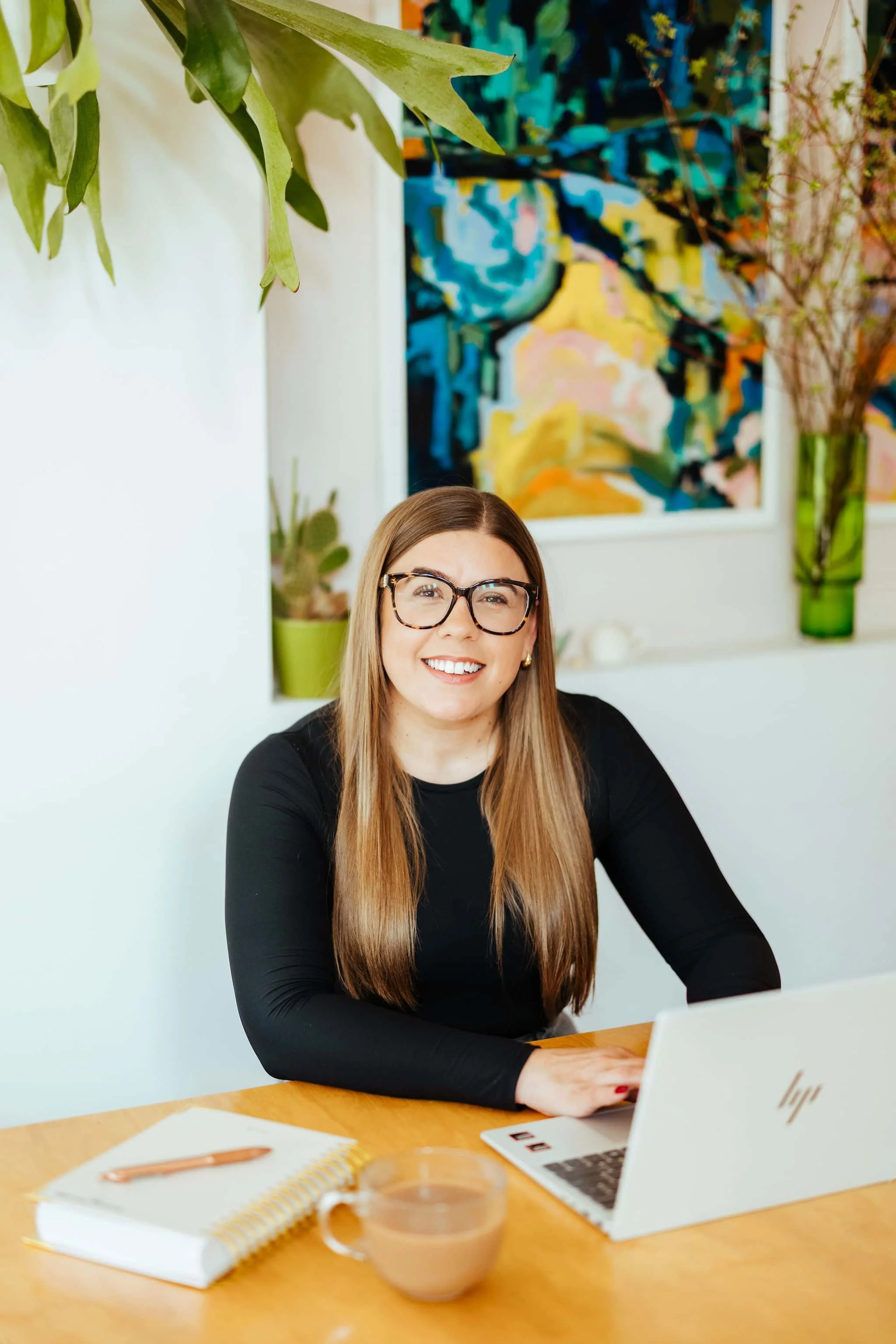 A woman with long brown hair, glasses, and a black top smiling while sitting at a wooden desk with a laptop, notebook, pen, and a cup of coffee, in a room with plants and a colorful abstract painting on the wall.