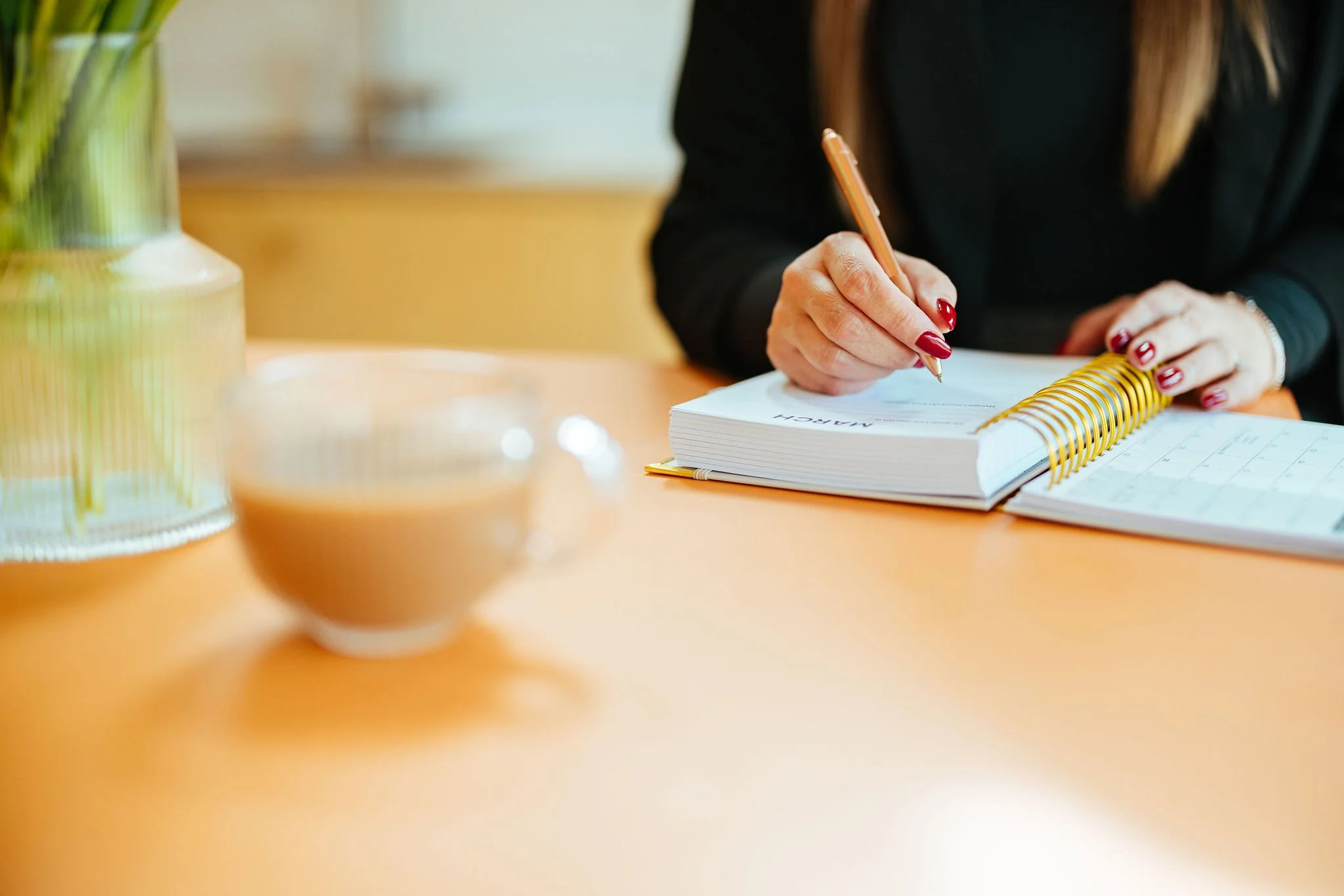 A woman with red painted nails writing in a spiral-bound planner or journal on an orange table, with a blurred cup and a green vase nearby.