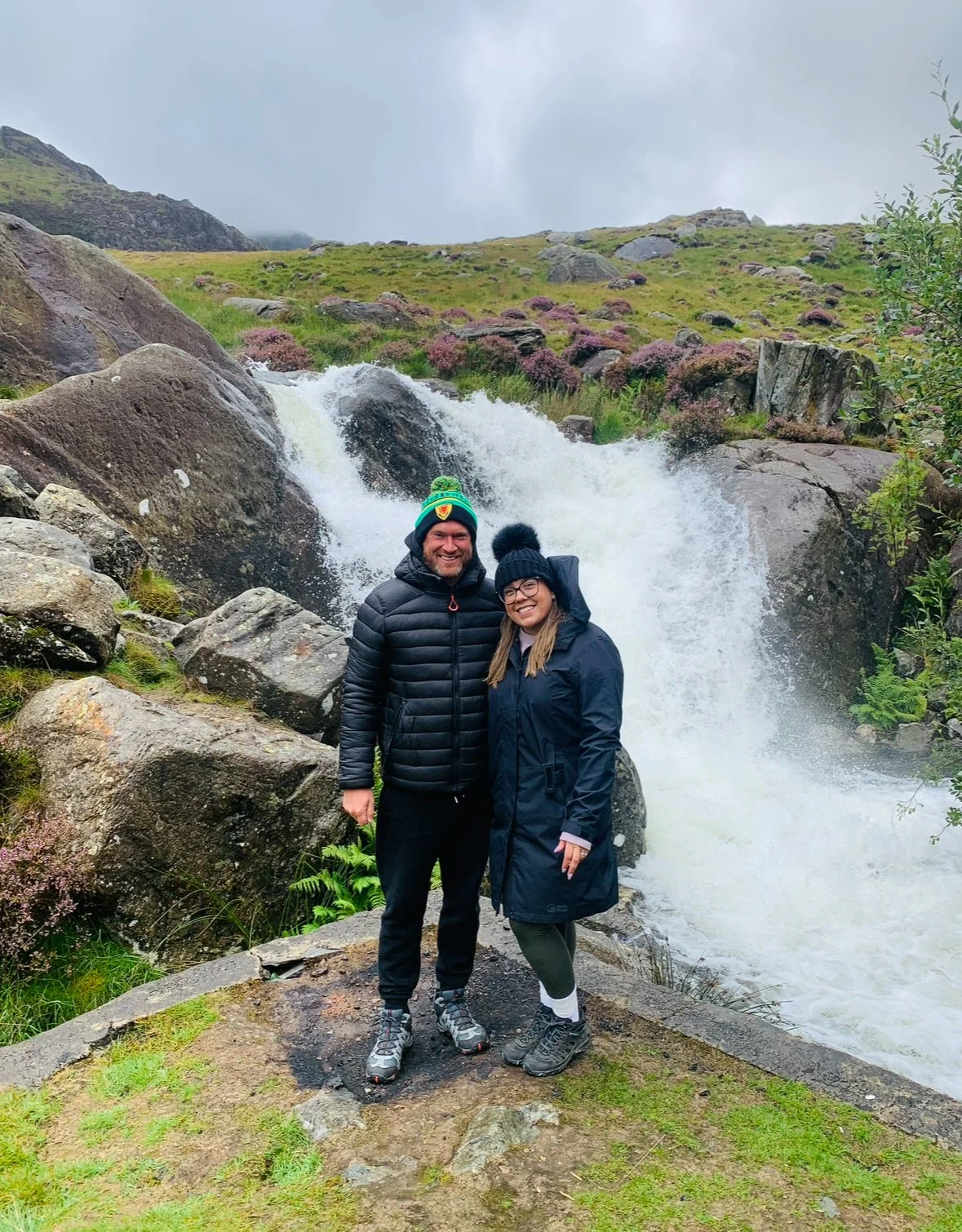 Two people smiling and standing together outdoors in front of a waterfall, wearing warm winter clothing, including hats and jackets.
