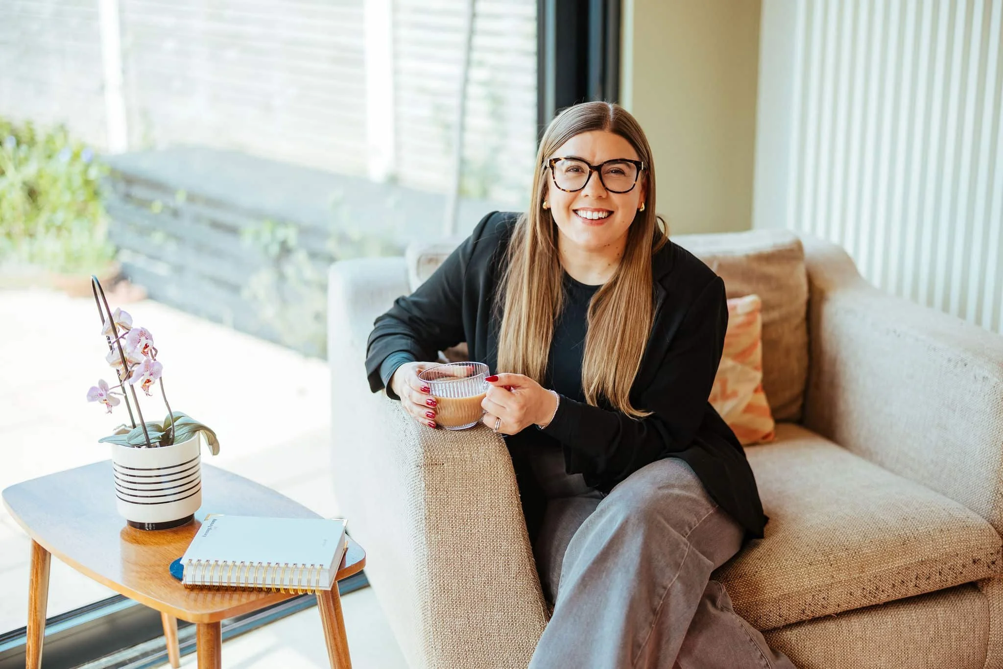 A woman with long hair, glasses, and a black blazer sitting on a beige sofa, holding a mug, smiling in a living room with a coffee table, potted orchid, and open window.