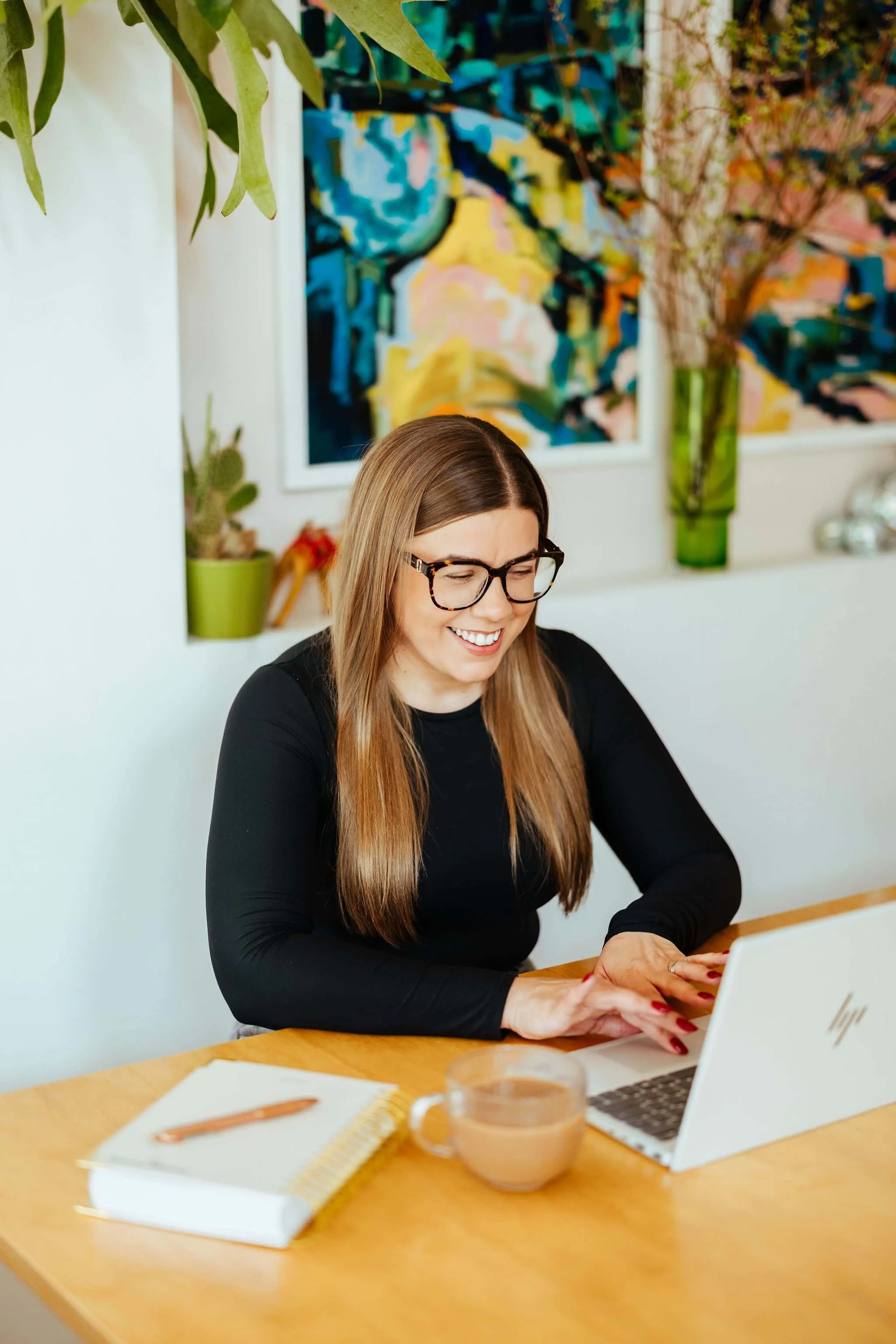 A woman with long brown hair and glasses smiling while working on a laptop at a wooden desk, surrounded by colorful artwork and potted plants in a bright room.