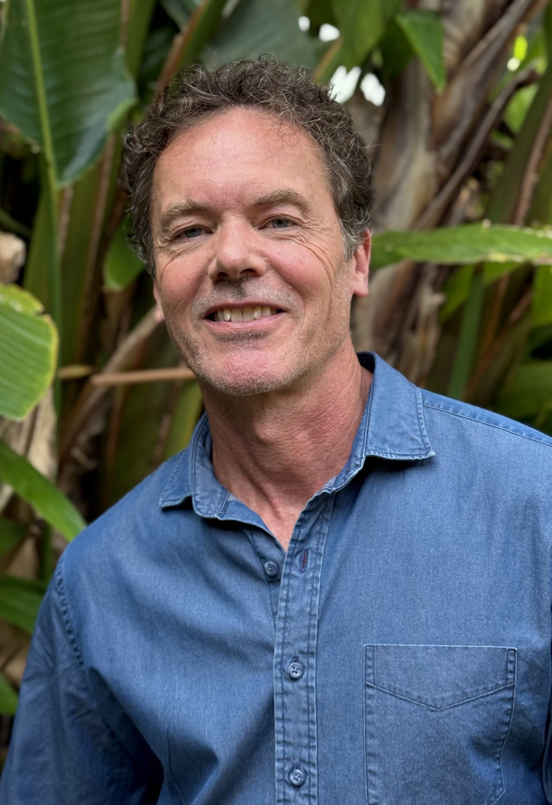 A middle-aged man with curly, dark hair and a light beard, smiling in front of lush green tropical plants, wearing a blue denim shirt.