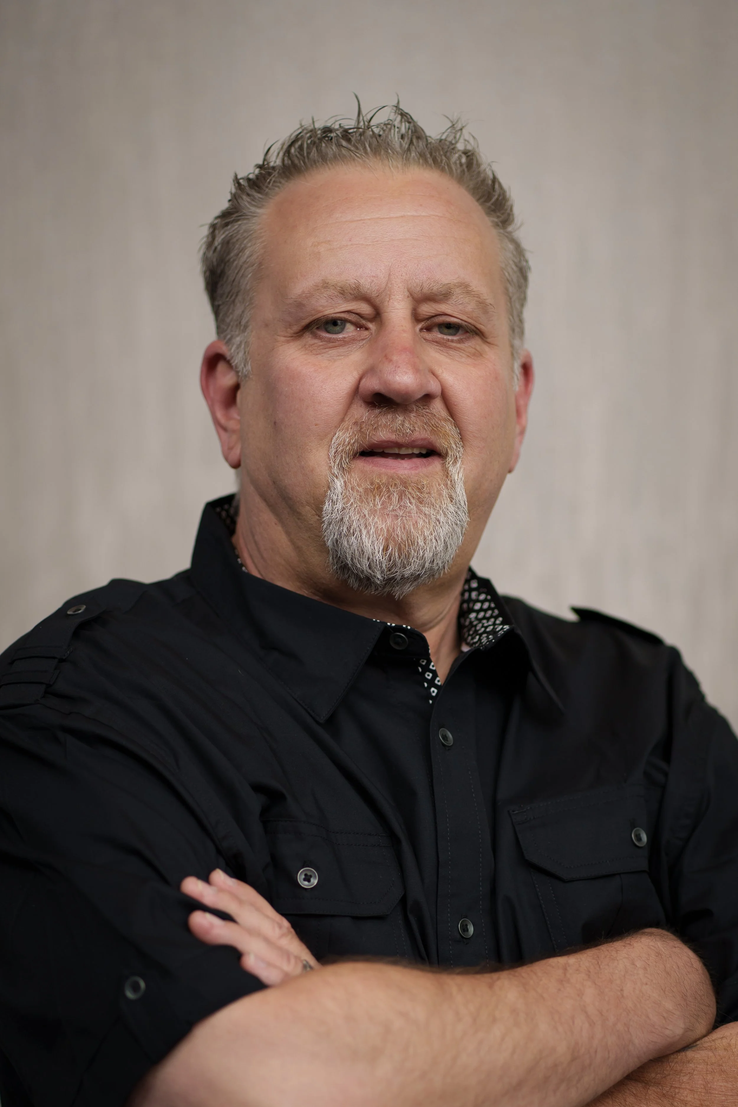 A middle-aged man with gray hair and a beard, wearing a black shirt with a patterned collar, is standing with arms crossed in front of a plain, light-colored background.