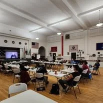 A large conference room with round tables and chairs, some people seated, a big screen TV on the wall, and American flags in the corners.