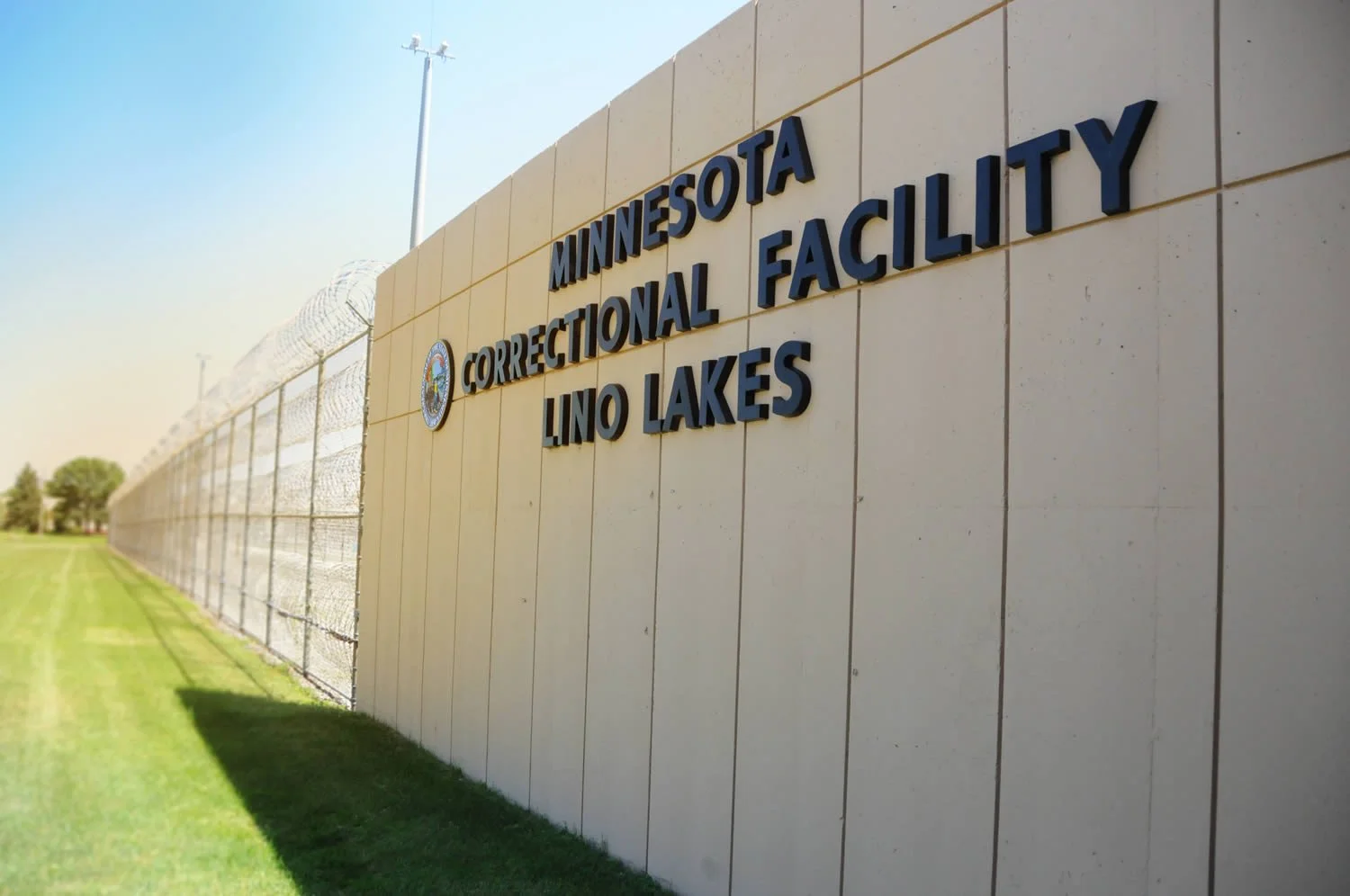 Exterior of the Minnesota Correctional Facility in Lino Lakes, showing a tall chain-link fence topped with barbed wire, and a beige wall with a sign.