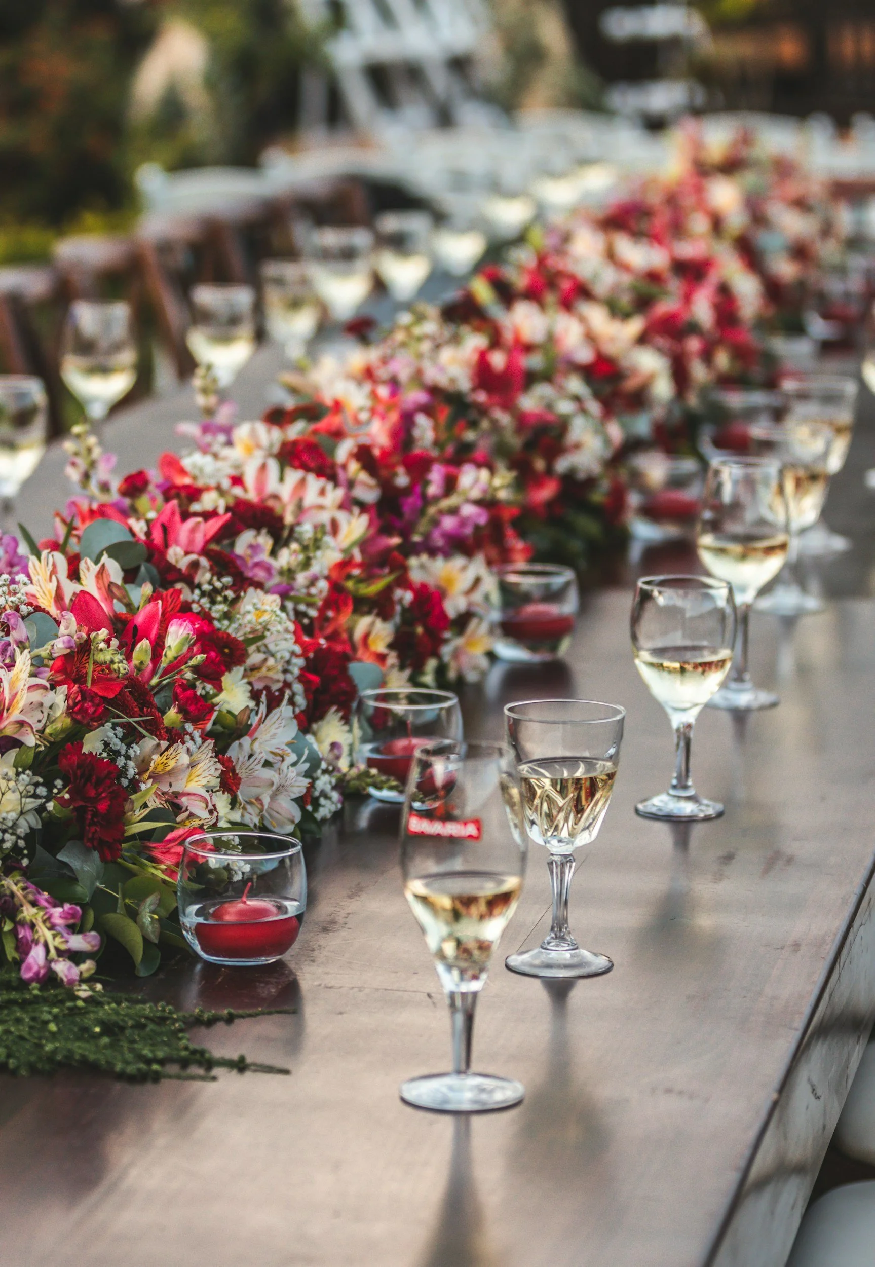 Long table with floral centerpiece and glasses of white wine, set for a celebration or gathering, outdoors on a sunny day.