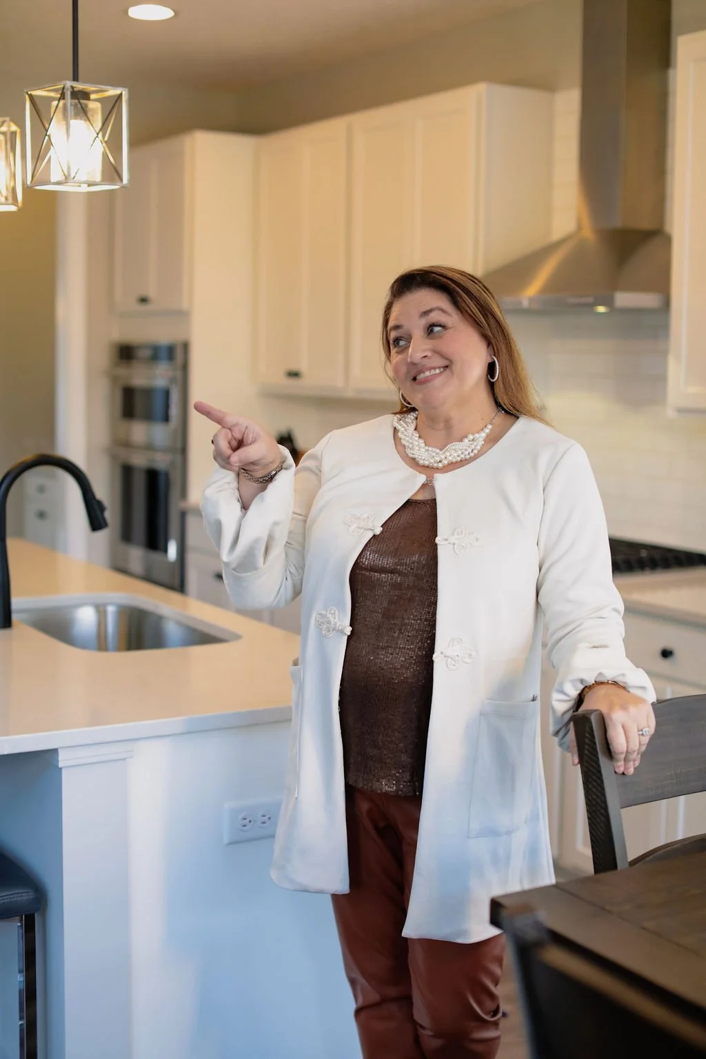 A woman standing in a kitchen, smiling and pointing to her left while holding a chair with her right hand. The kitchen has white cabinets and stainless steel appliances.