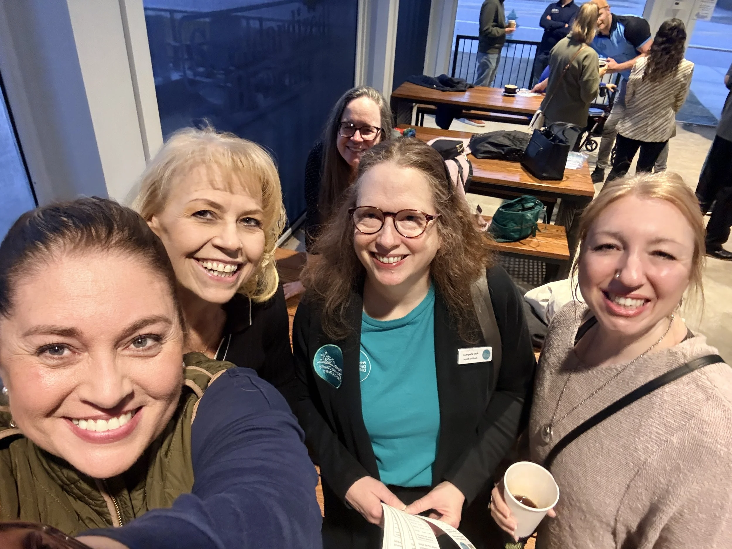 A group of people smiling for a selfie at a social gathering, with some holding cups of coffee or beverages inside a building with large windows.