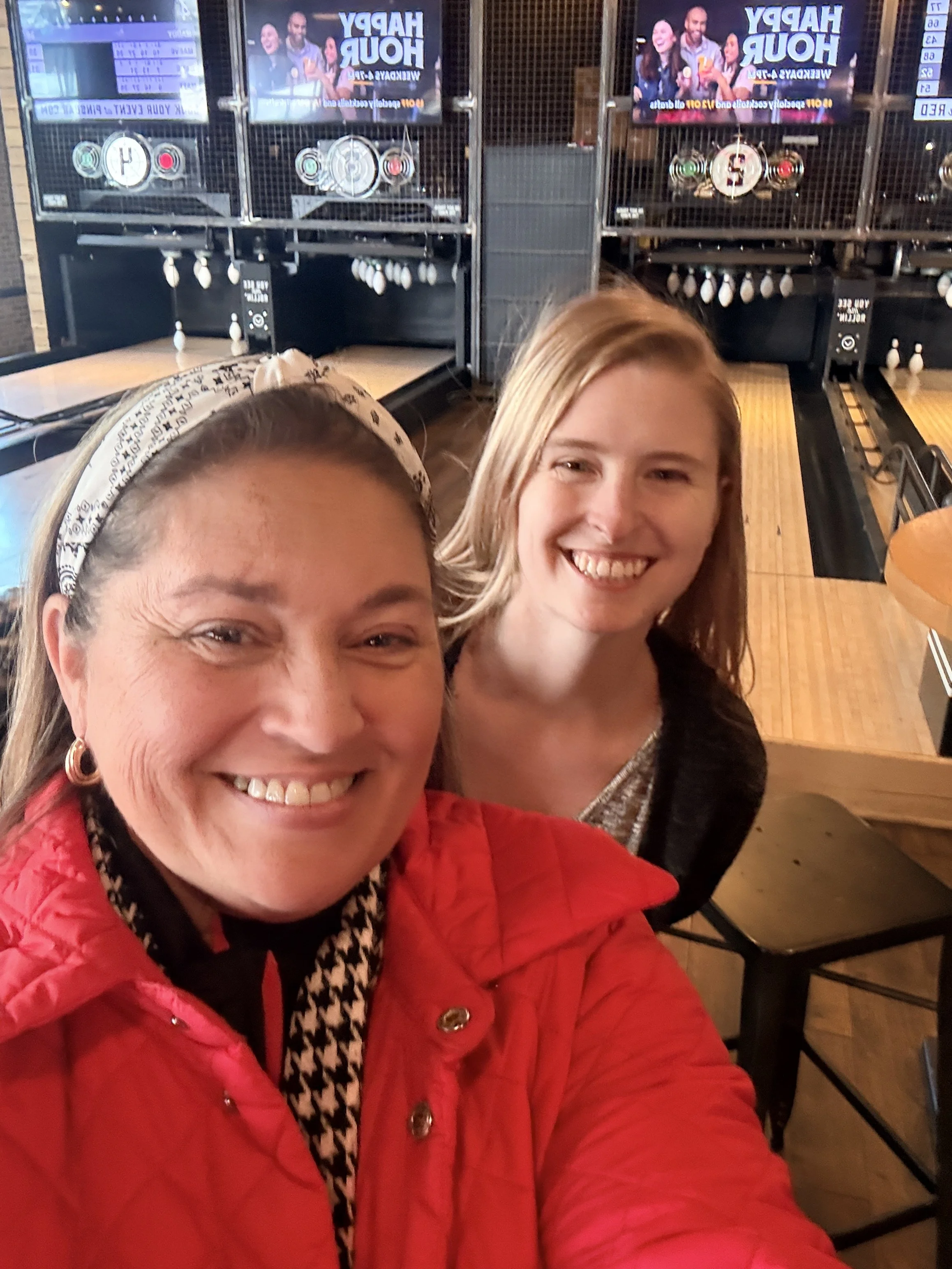 Two smiling women taking a selfie at a bowling alley, with bowling lanes and digital screens in the background.