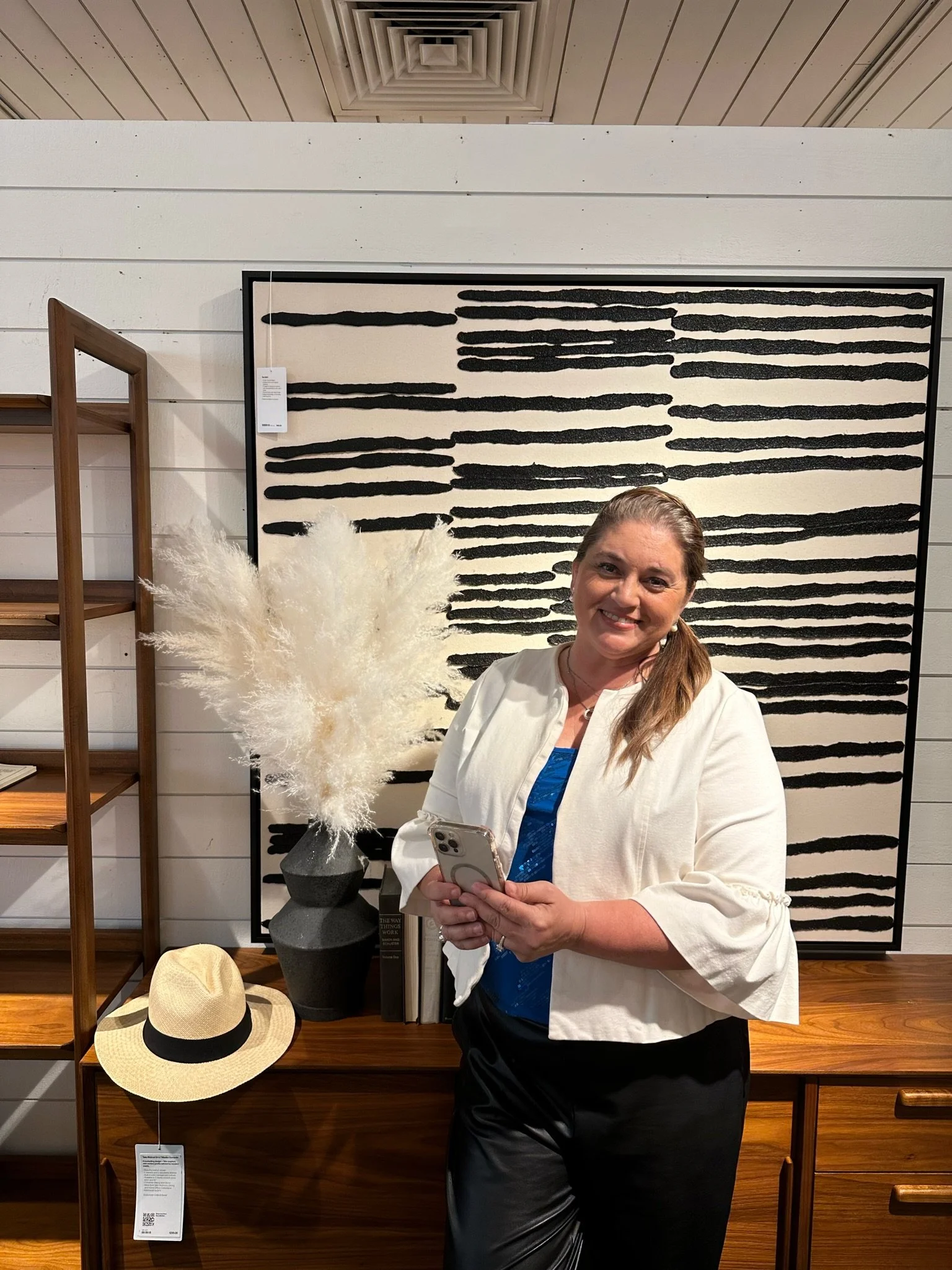 A woman with brown hair, dressed in a white blazer and black skirt, smiling and holding a phone, standing in front of a black and white abstract artwork in a retail store. There is a beige hat with a black band on the wooden table beside her, a tall 
