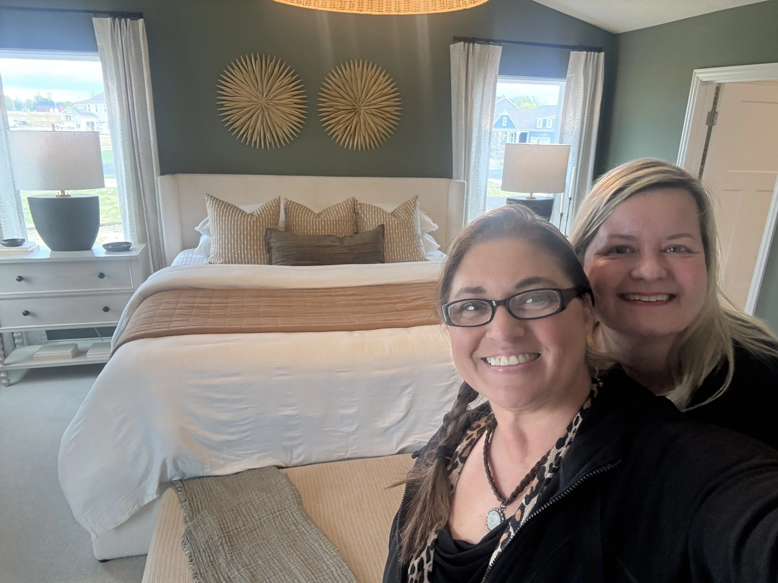 Two women smiling taking a selfie in a bedroom with a bed, decorative pillows, two nightstands with lamps, and artwork on the wall.