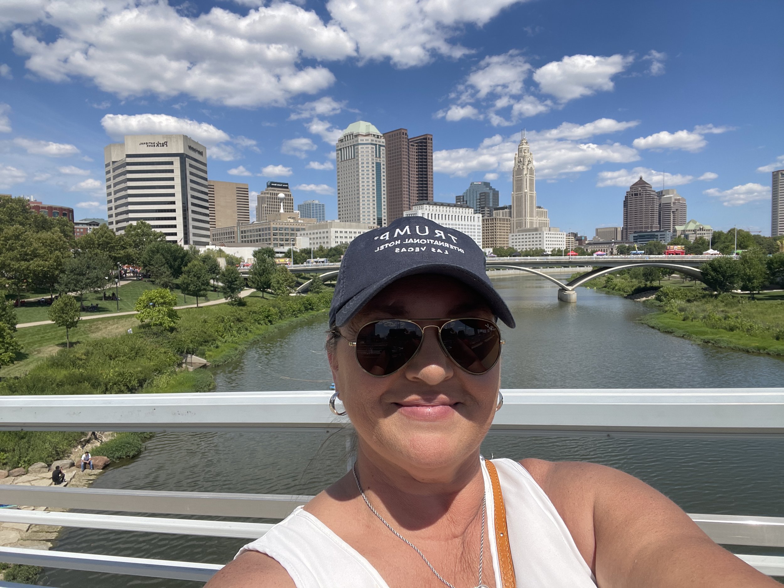 A woman with sunglasses and a navy baseball cap taking a selfie on a bridge overlooking a river, with a city skyline including skyscrapers and a church spire under a partly cloudy sky.