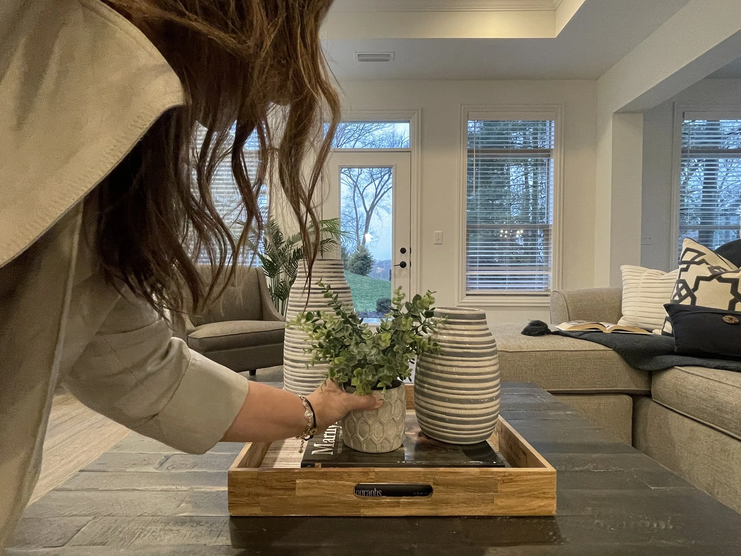 A woman with long brown hair arranging a potted plant on a wooden table in a living room with large windows and modern decor.