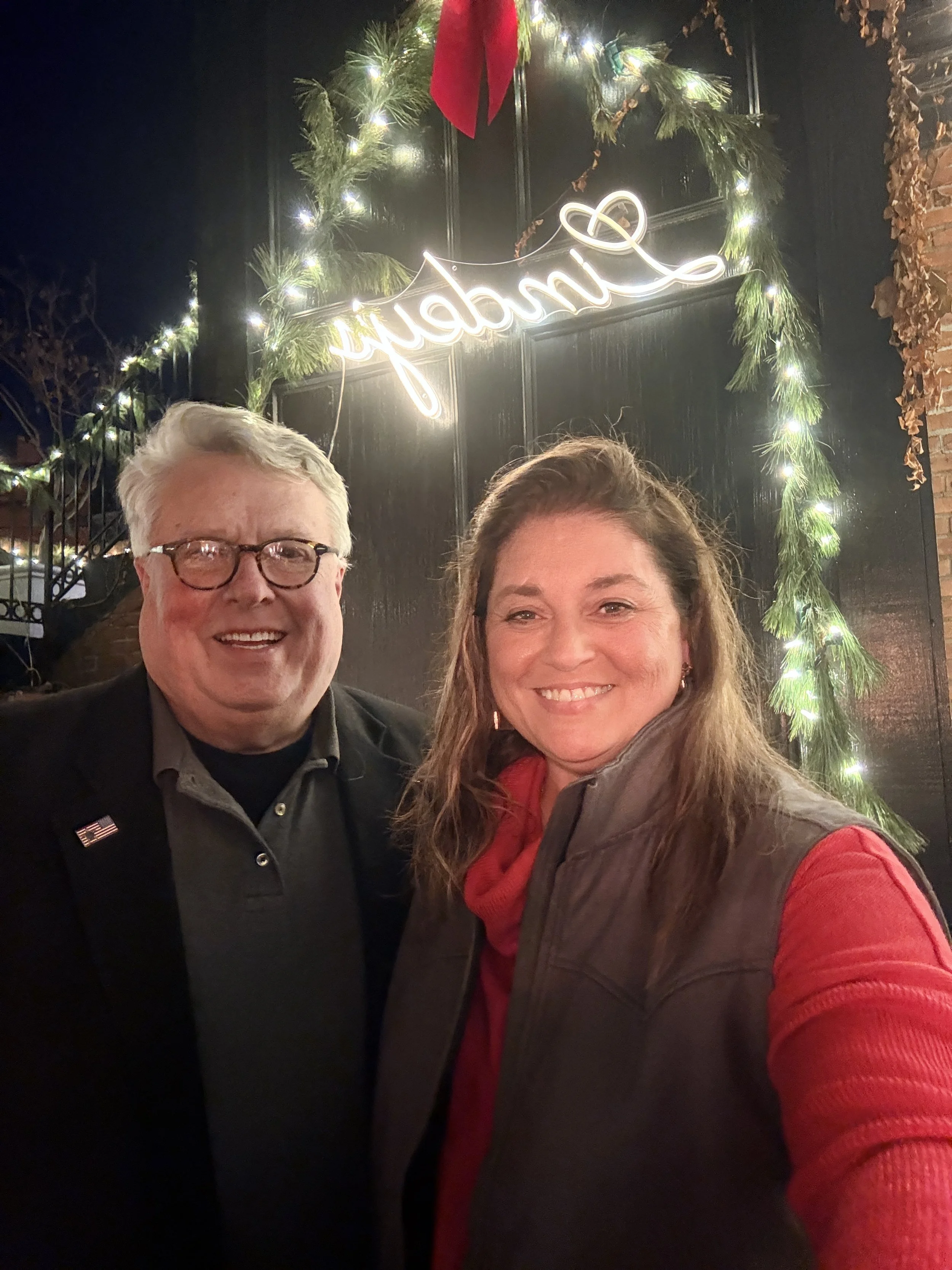 Two smiling people taking a selfie in front of a festive holiday decoration featuring a lit sign that says 'Jubilant', surrounded by greenery and Christmas lights at night.