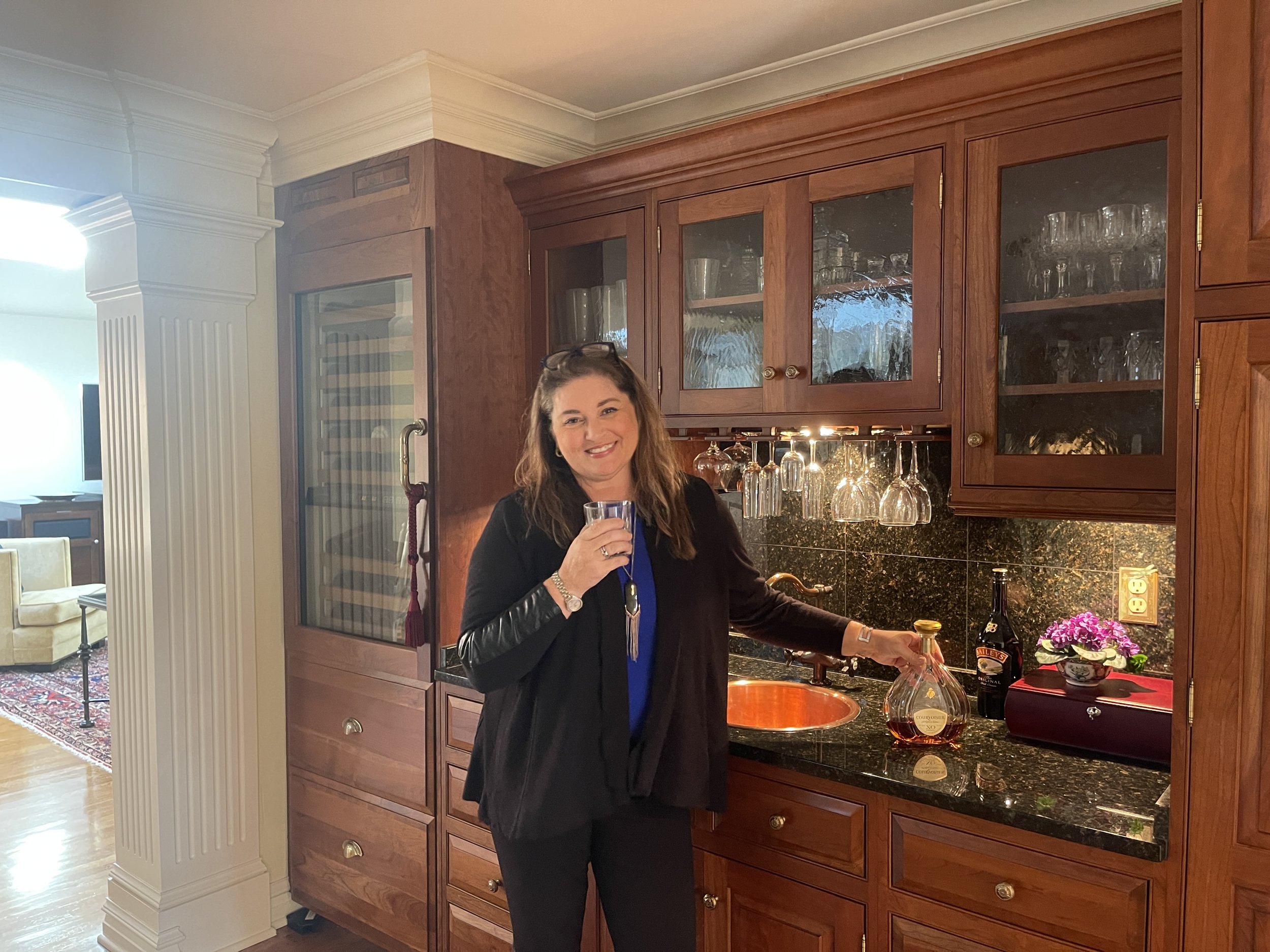 A woman smiling while holding a glass in a kitchen with wooden cabinets, black granite countertops, and wine glasses hanging upside down. There is a bottle of whiskey, a bouquet of pink and white flowers, and a wine bottle on the counter.