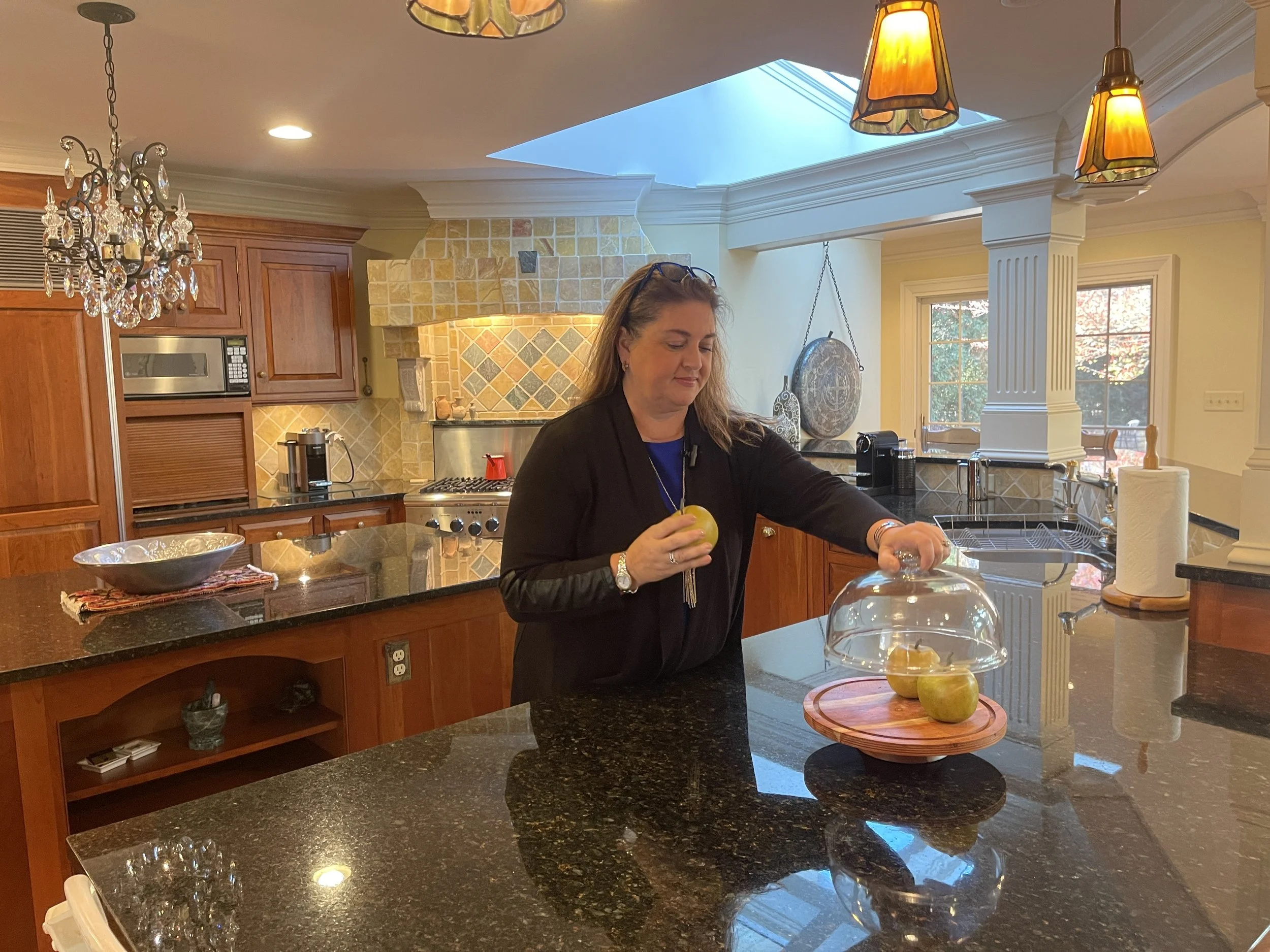 A woman in a black jacket is standing at a kitchen counter, holding a yellow apple. There are three apples on a round wooden tray in front of her. The kitchen features wooden cabinets, a granite countertop, a mosaic tile backsplash, a chandelier, and