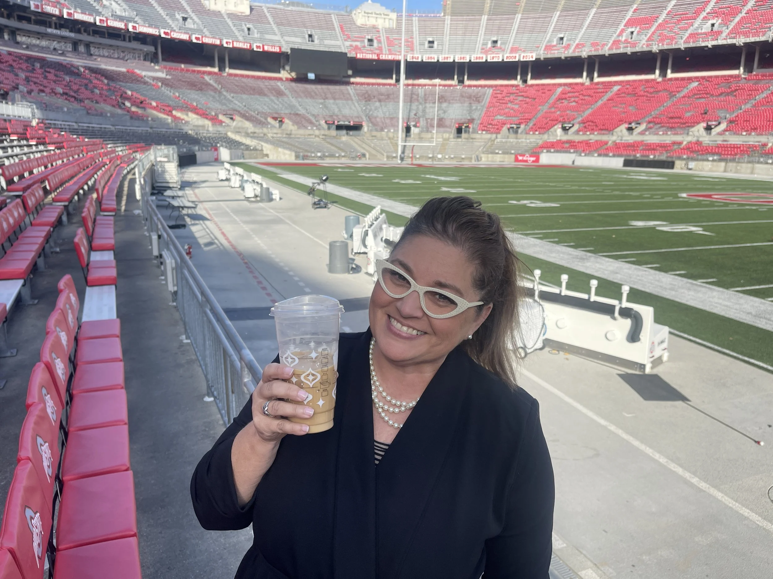 Smiling woman with glasses, Pearl necklace, and dark attire holding a large iced coffee in a stadium seating area with red seats and a football field in the background.