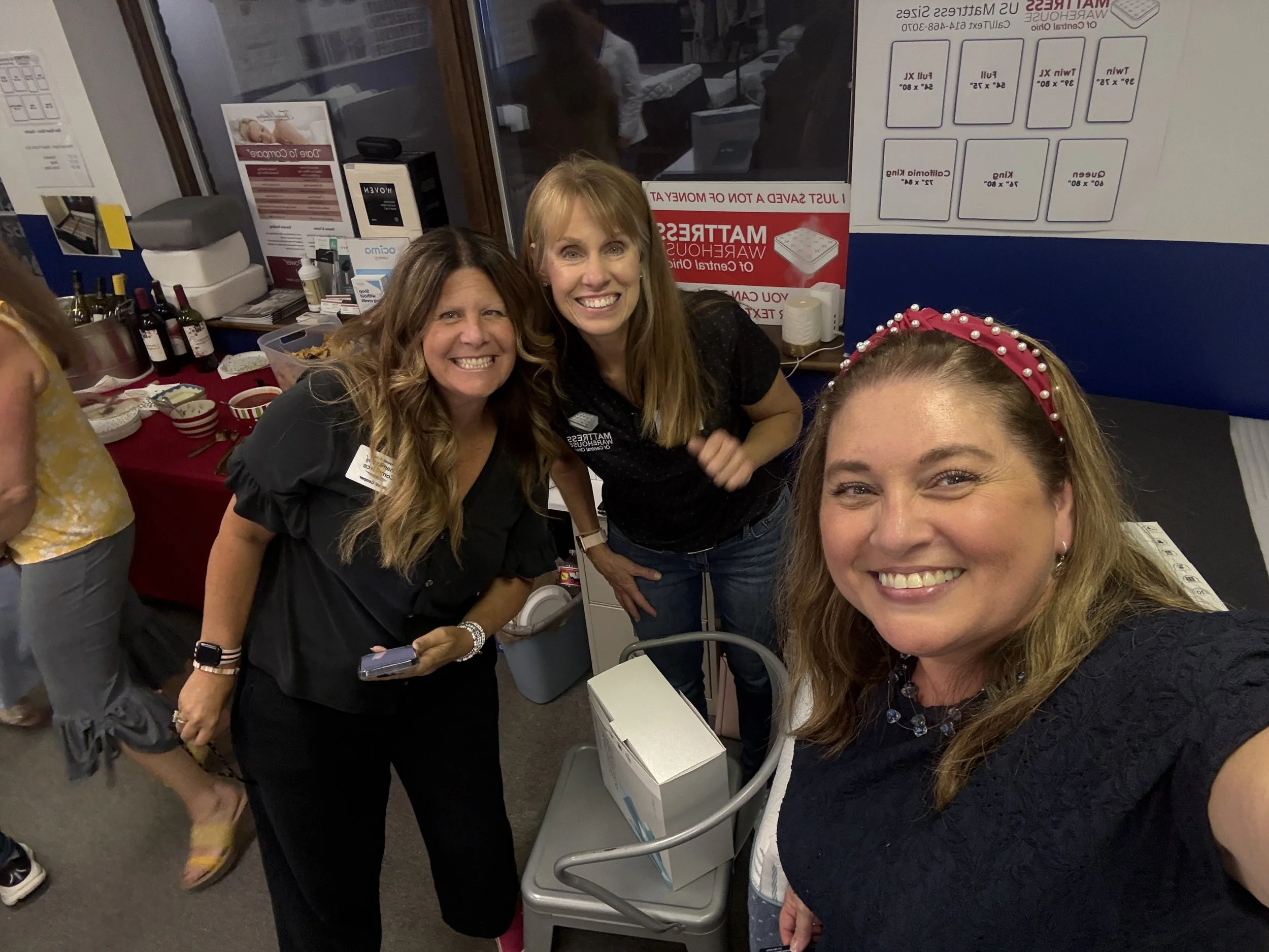 Group of women smiling indoors, some with name tags, a table with snacks and beverages, posters on the wall, and a window showing the room behind.
