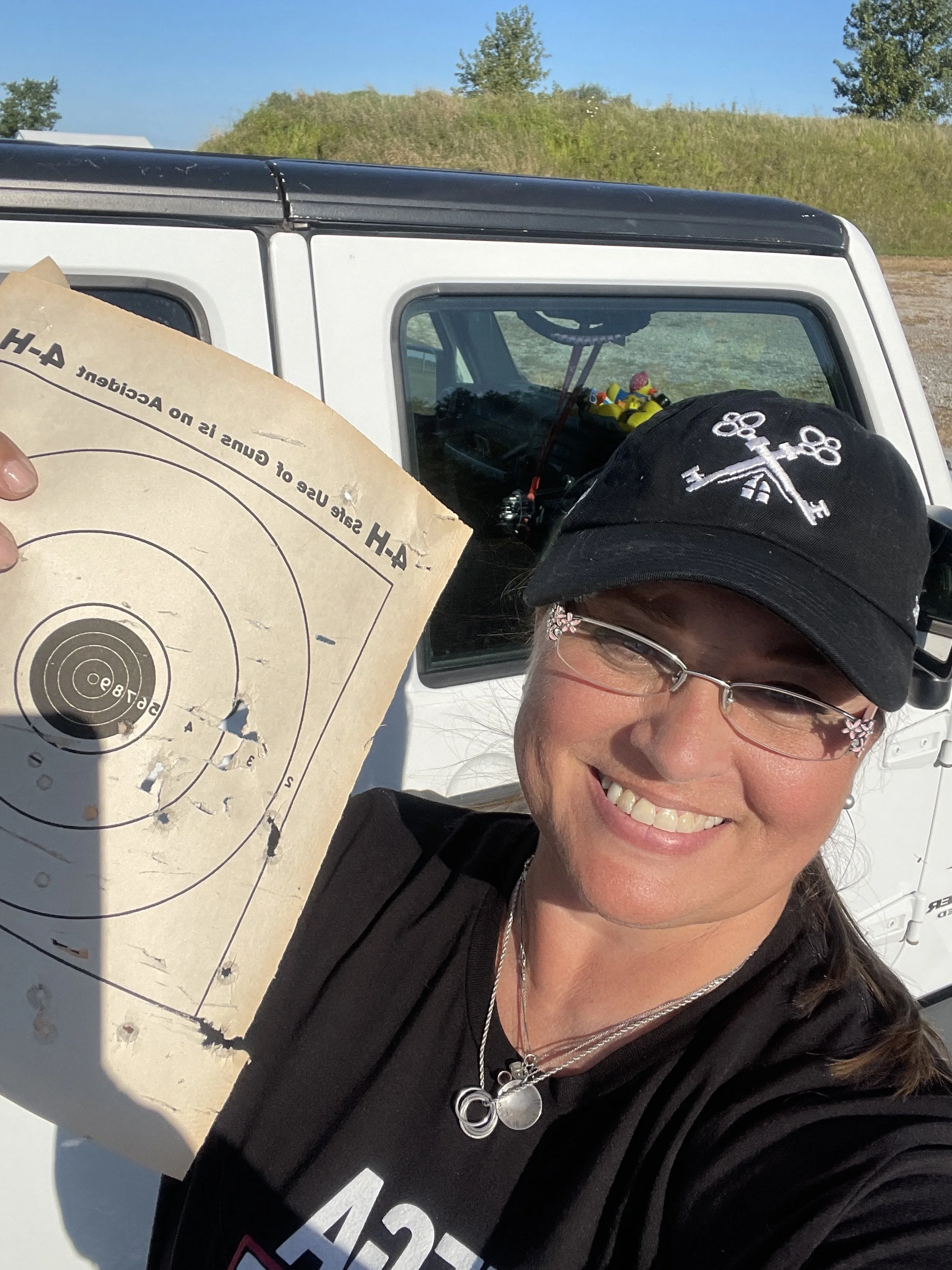Smiling woman with glasses and necklace holding a paper target at an outdoor shooting range with a white van in the background.