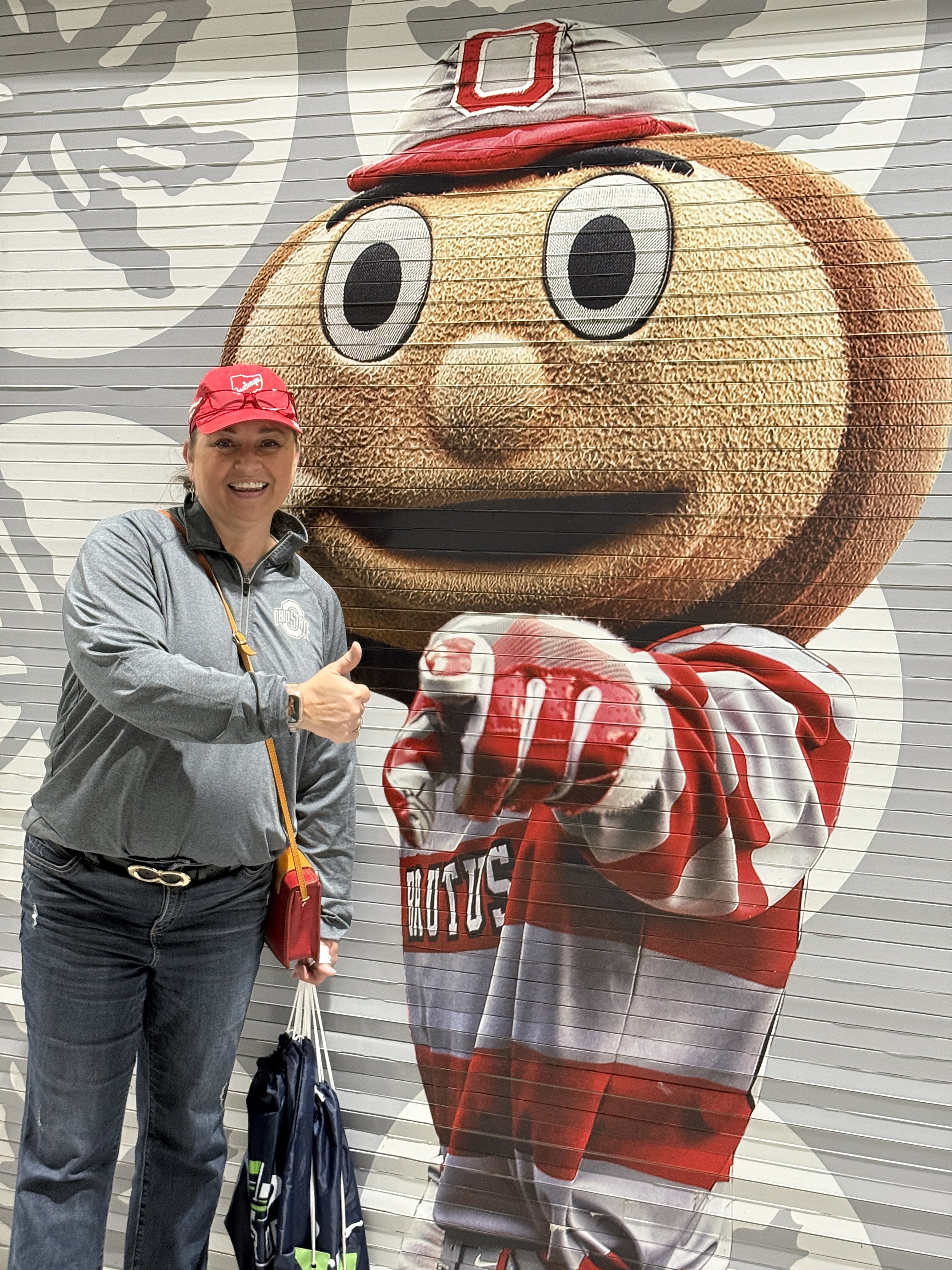 A woman smiling and giving a thumbs up standing in front of a large Michigan football mascot image on a wall.