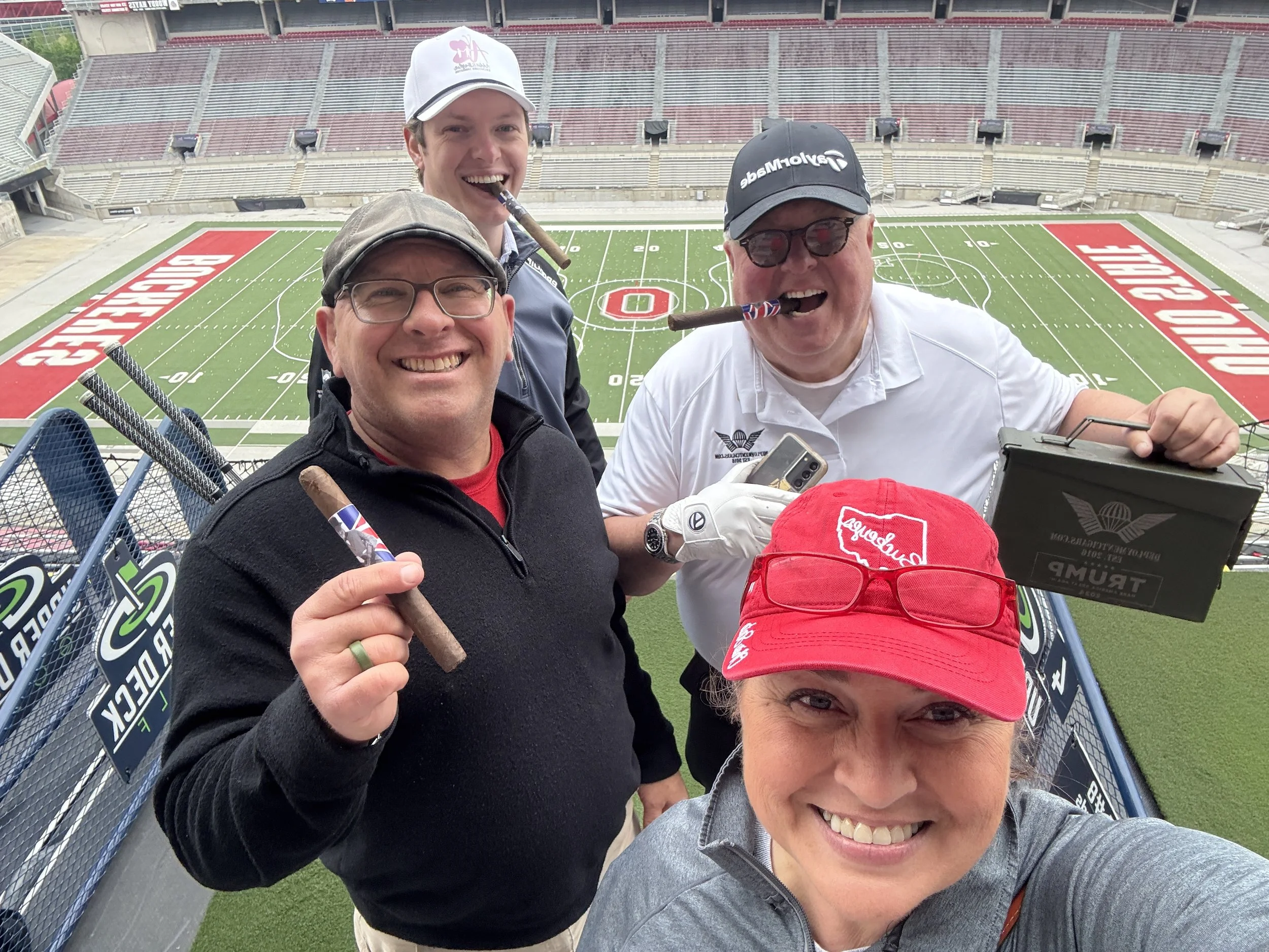 Four smiling people on a sports stadium tier, holding cigars and taking a selfie. The football field below says 'Ohio State' with red end zones. They are wearing casual clothes and hats.