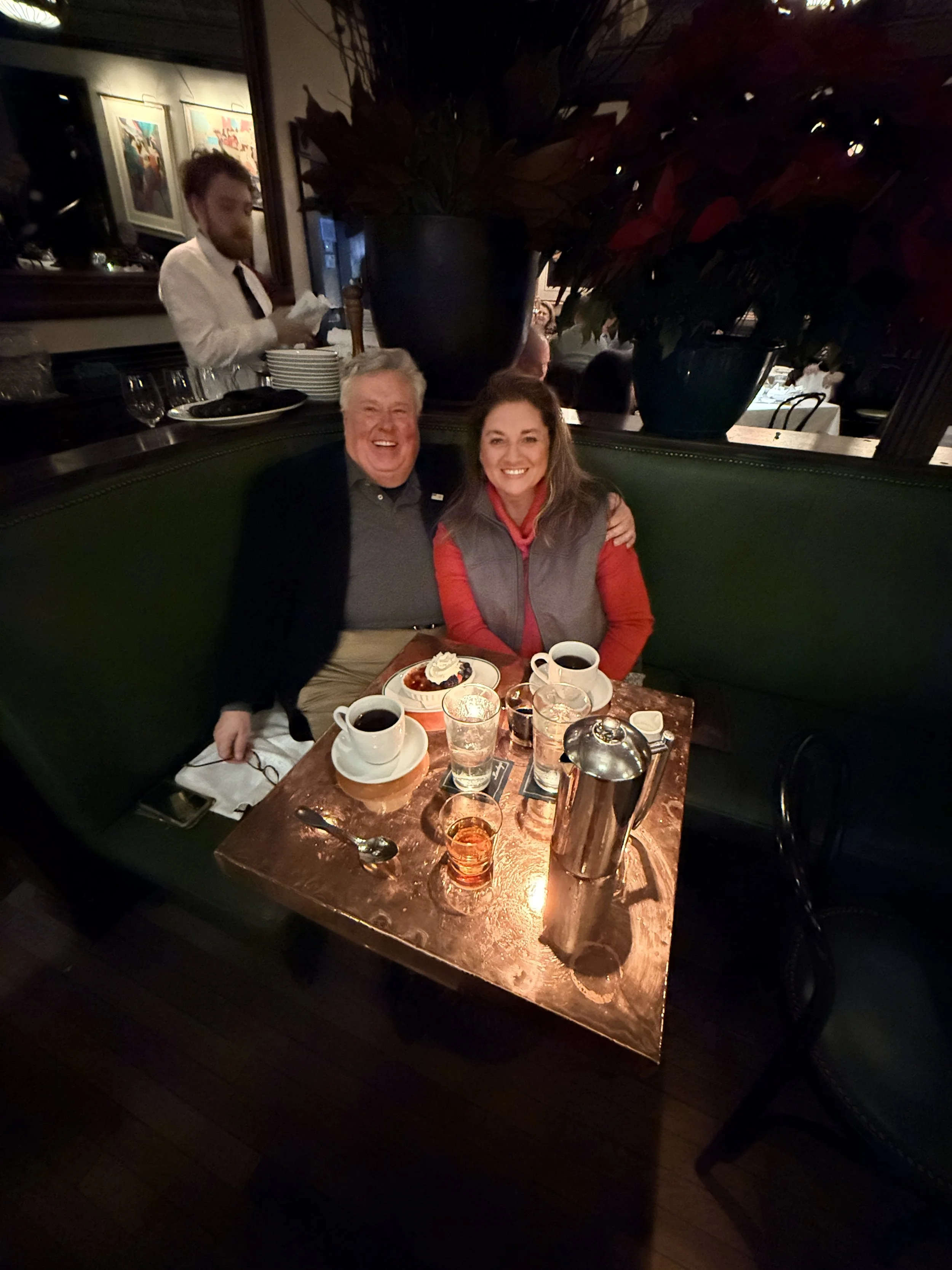 Two joyful people, a man and a woman, are sitting at a table in a dimly lit restaurant or cafe. The table has coffee cups, a small cake with whipped cream, glasses, and a metal ice bucket. There are large vases with flowers behind them. In the backgr