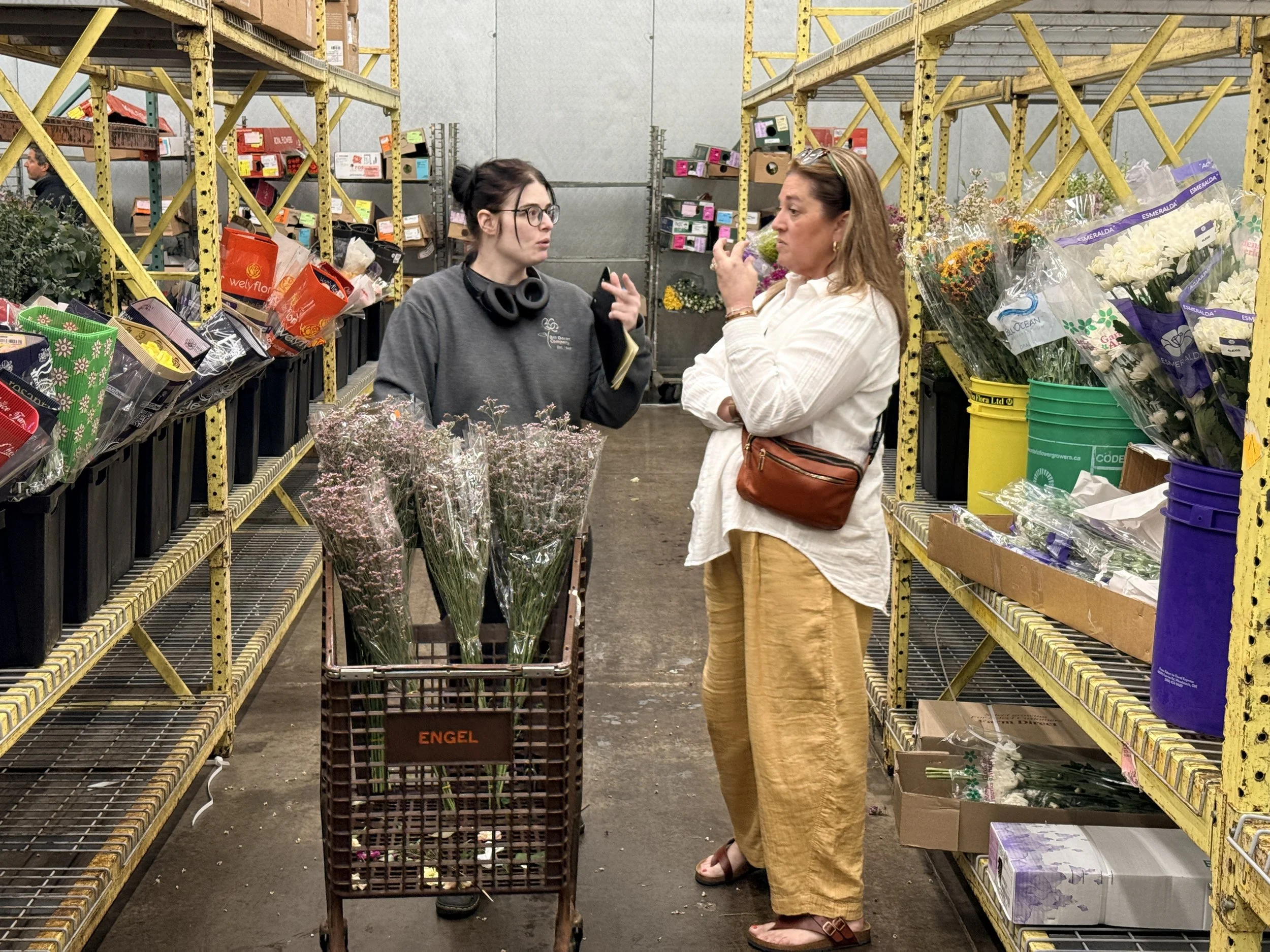 Two women are shopping for flowers in a warehouse-style store, surrounded by shelves stocked with flowers and floral arrangements. One woman is holding a small bouquet and talking to the other, who has her arms crossed.